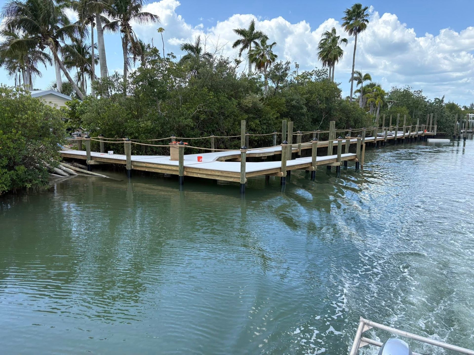 Mangrove-lined canal with wooden dock, calm blue water, and palm trees under a partly cloudy sky