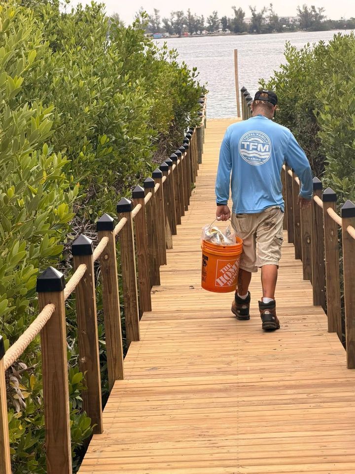 Person carrying an orange bucket walks down a wooden boardwalk between green mangroves toward water.
