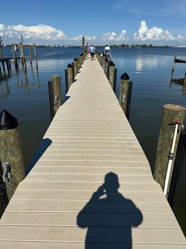 Wooden pier leading to calm water under a bright blue sky, with a person’s shadow in the foreground.