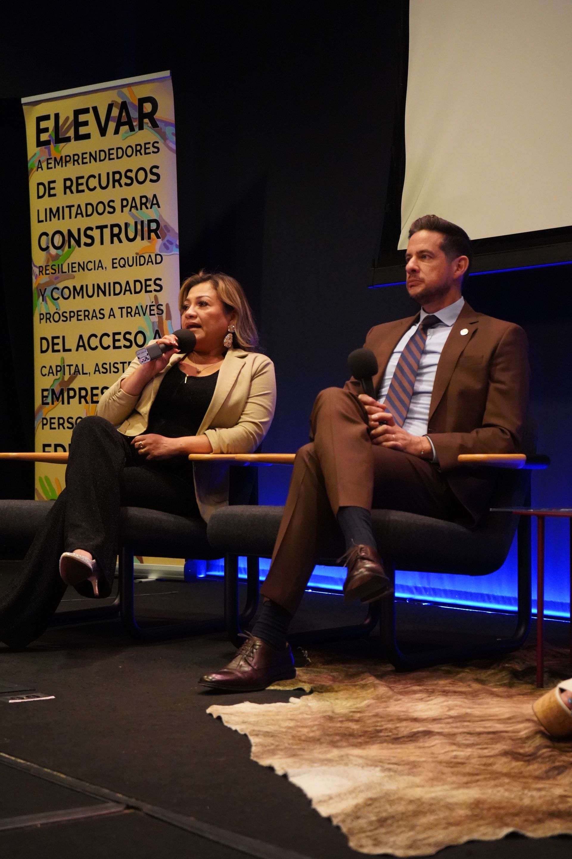 A man and a woman are sitting on a couch talking into microphones.