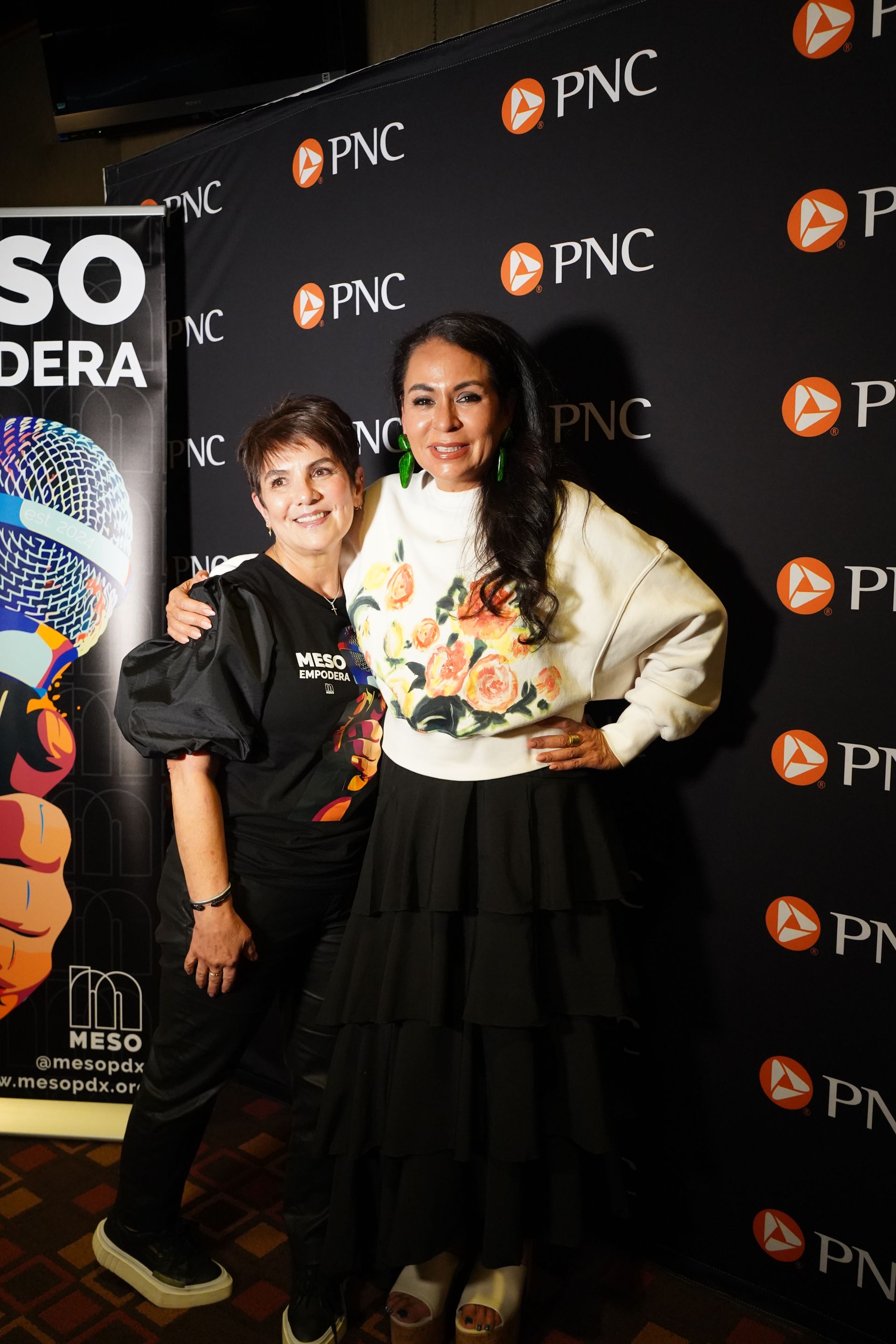 Two women are posing for a picture in front of a black wall.