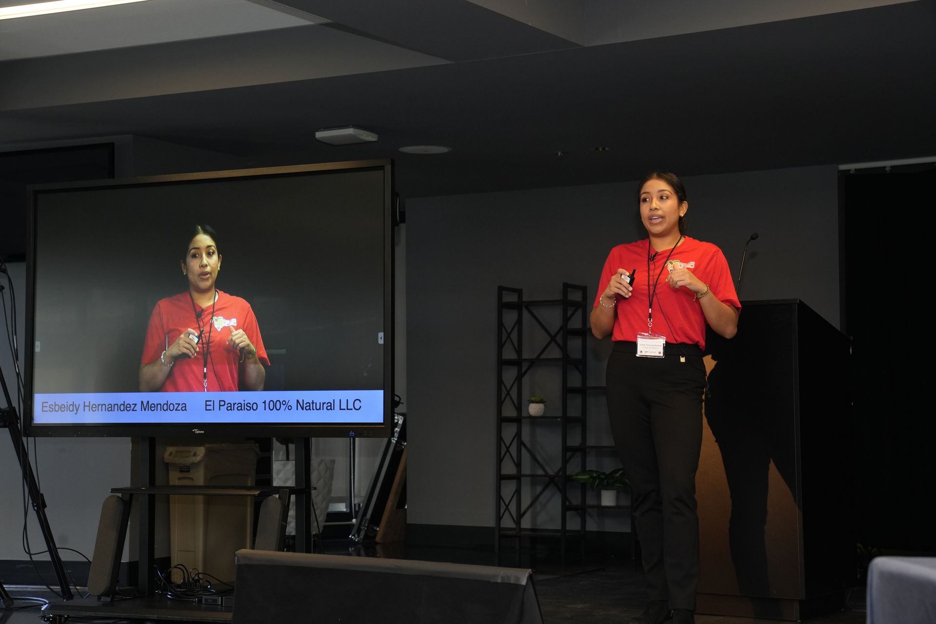 A woman in a red shirt is giving a presentation in front of a large screen.