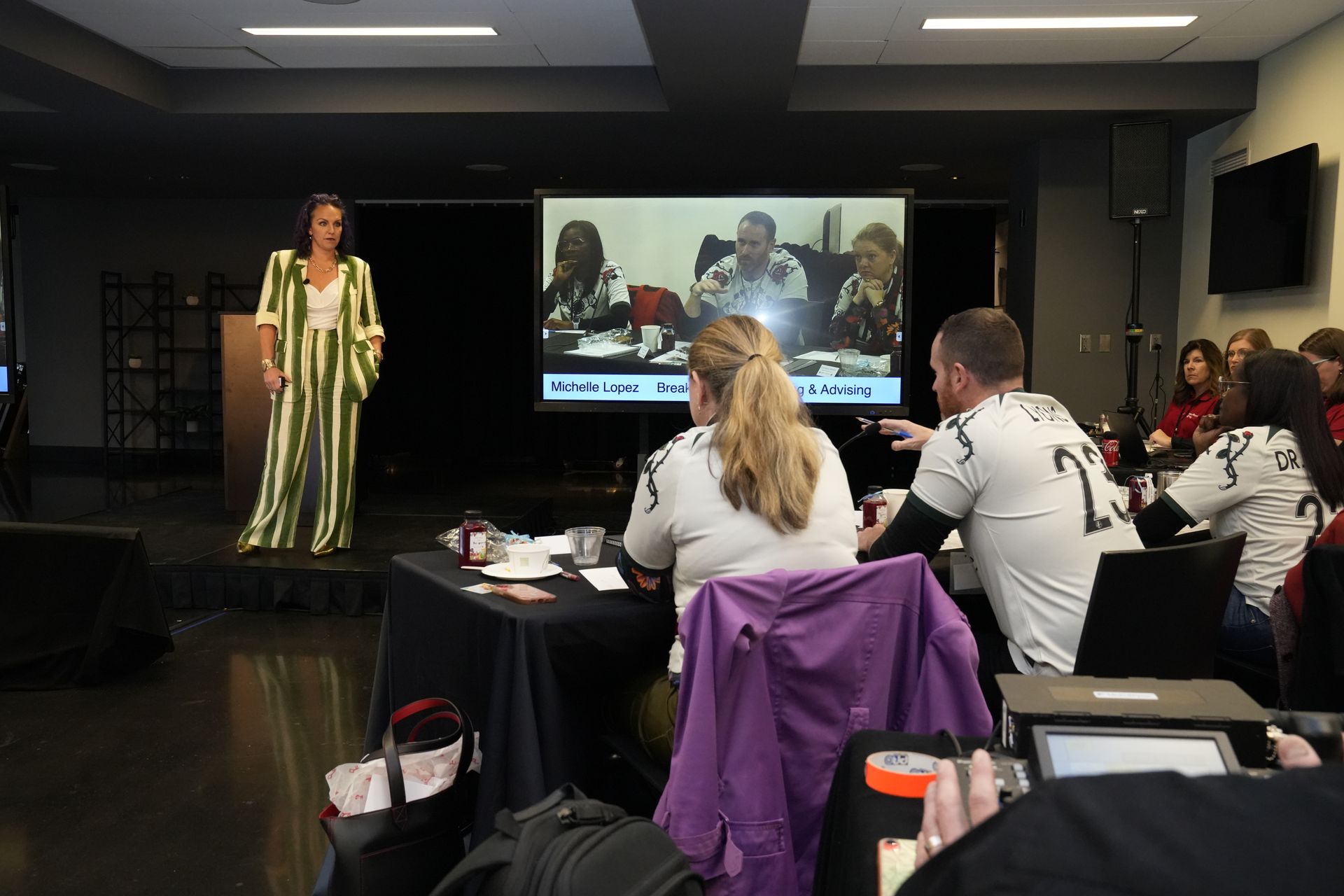 A woman is standing on a stage giving a presentation to a group of people sitting at tables.