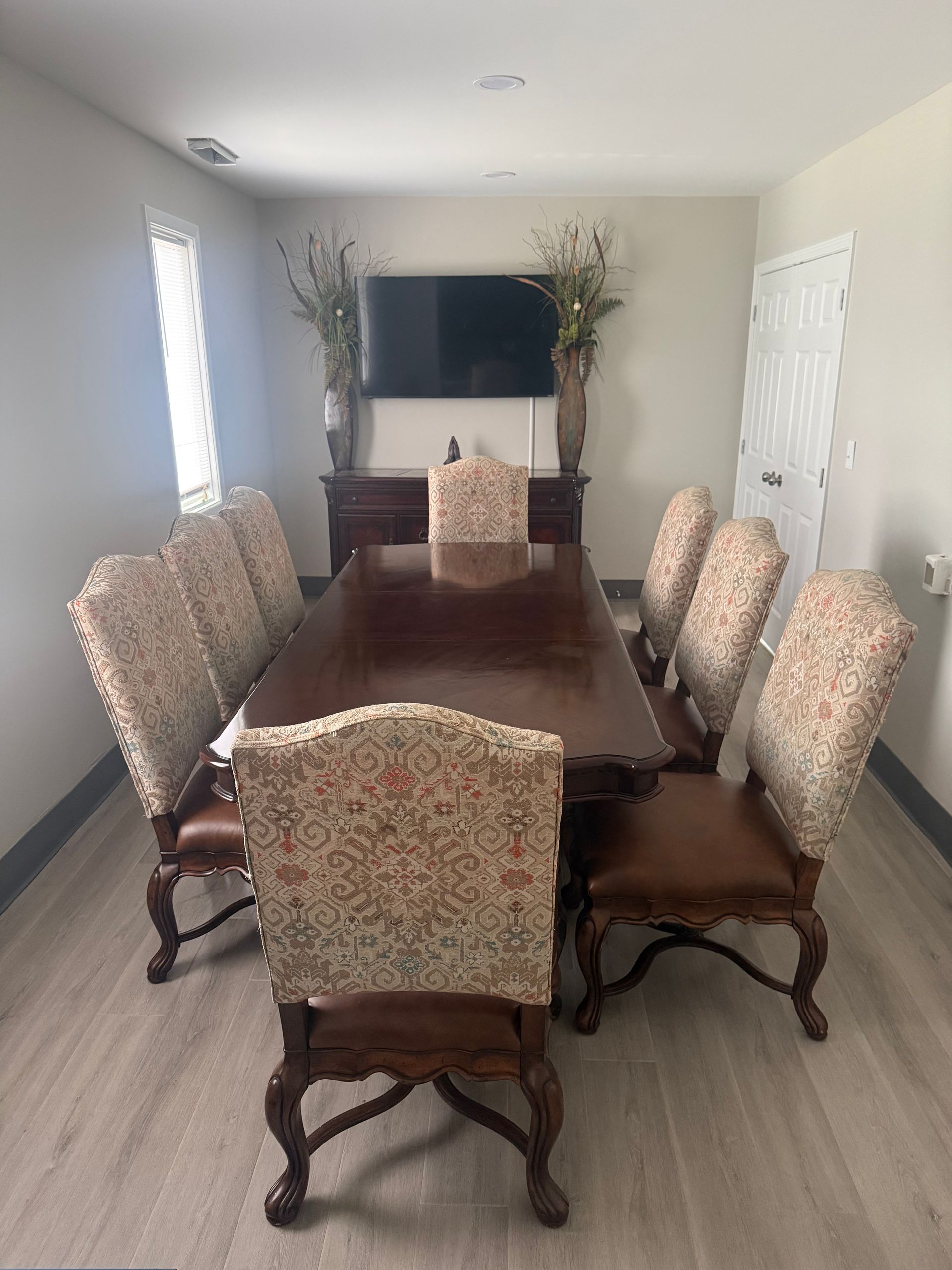 Dining room with a long wooden table and patterned chairs facing a TV and decorative vases.