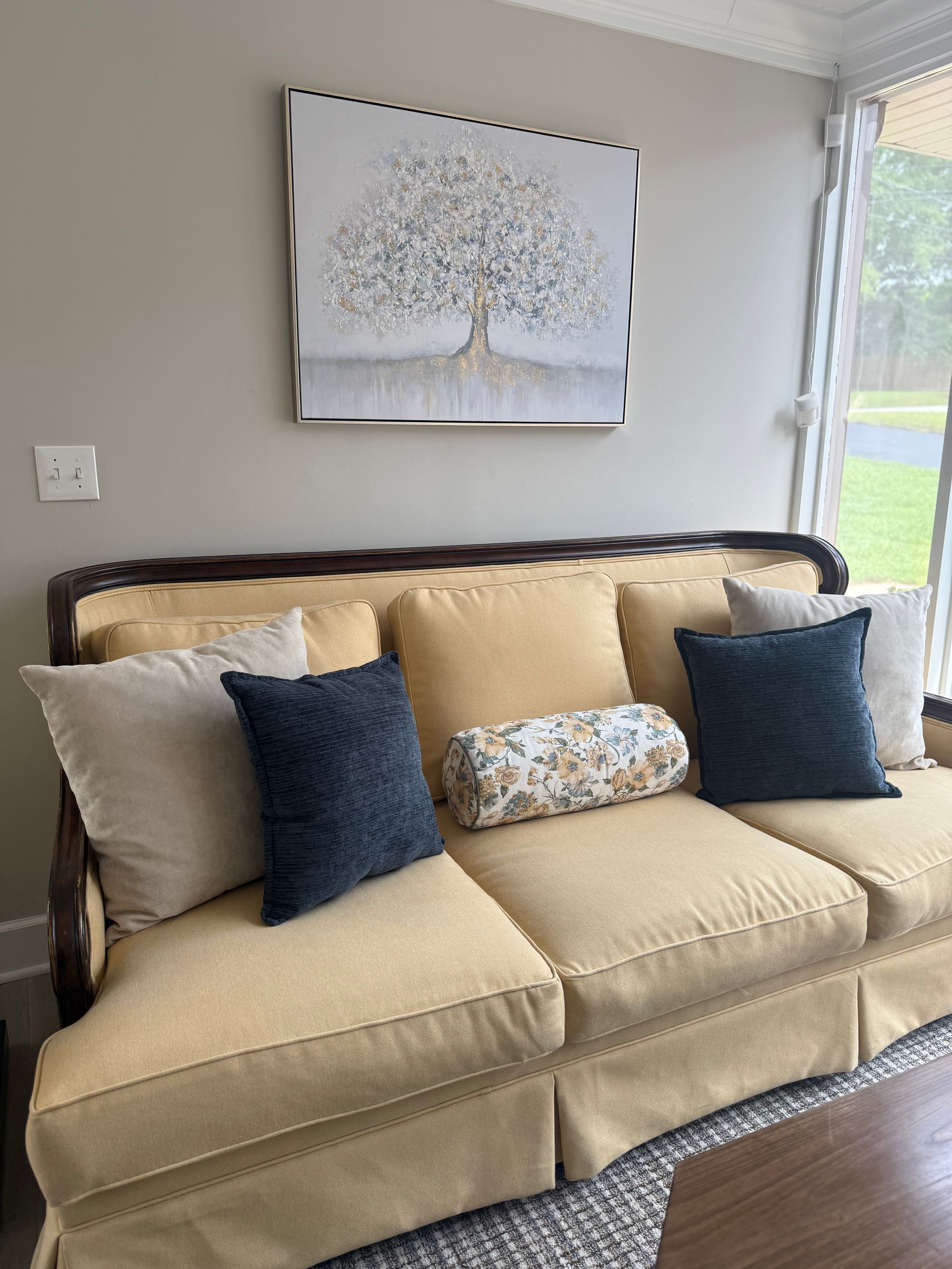 Beige sofa with blue and tan pillows beneath a tree wall art in a bright living room
