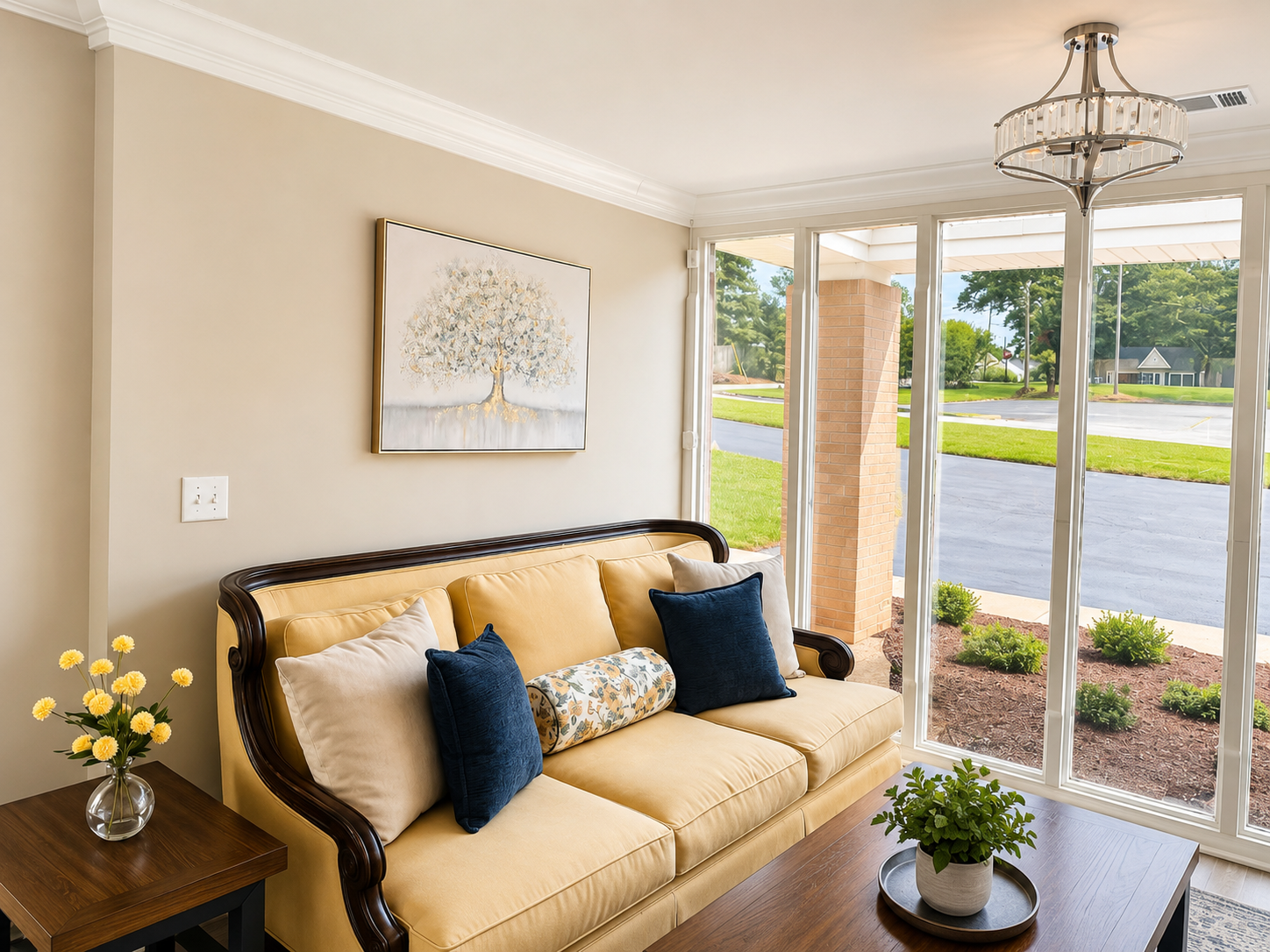 Cozy beige living room with sofa, decorative pillows, side table, and large front windows overlooking a street