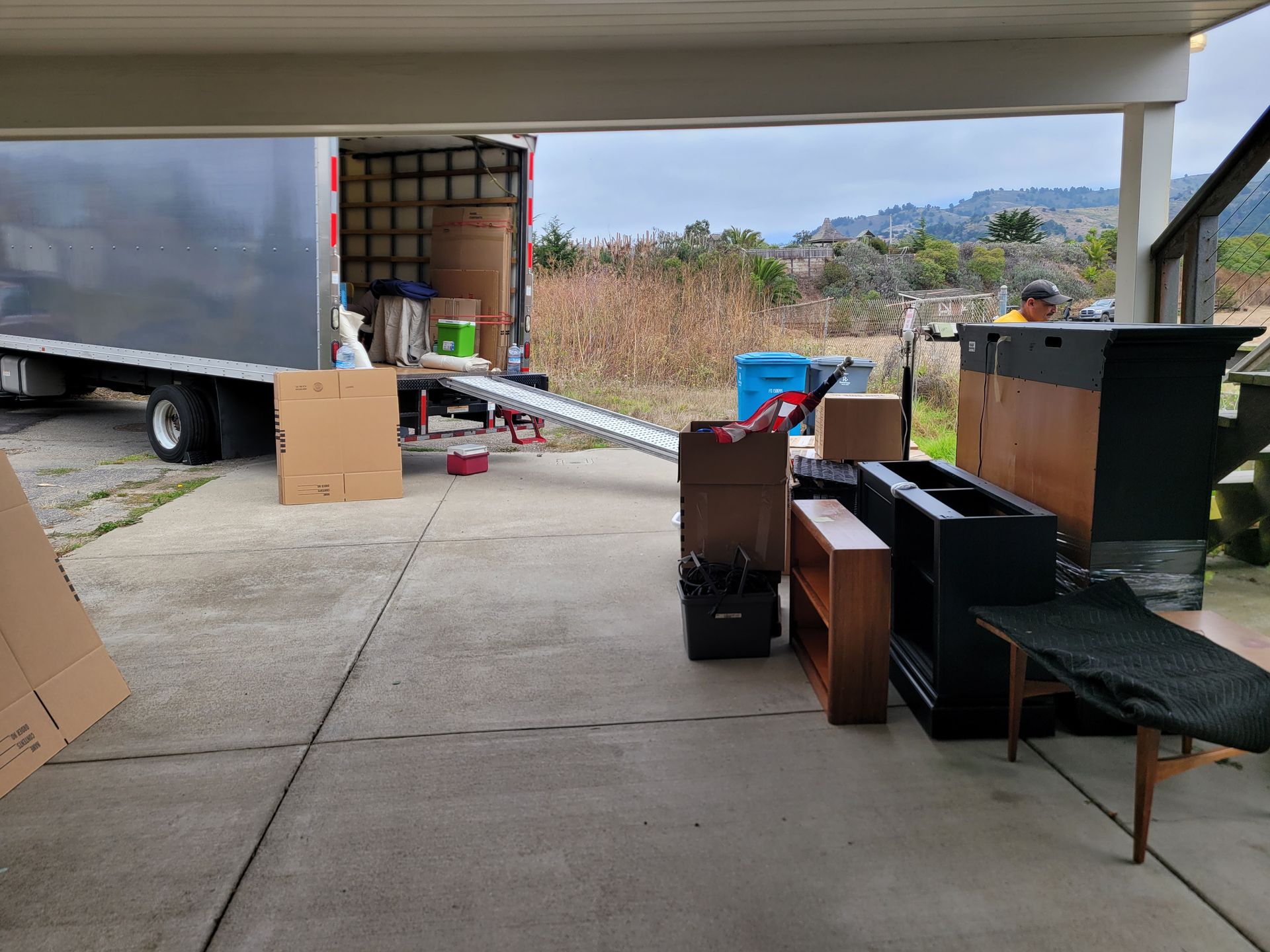 Covered outdoor area with a moving truck, boxes, and assorted furniture and appliances being unloaded