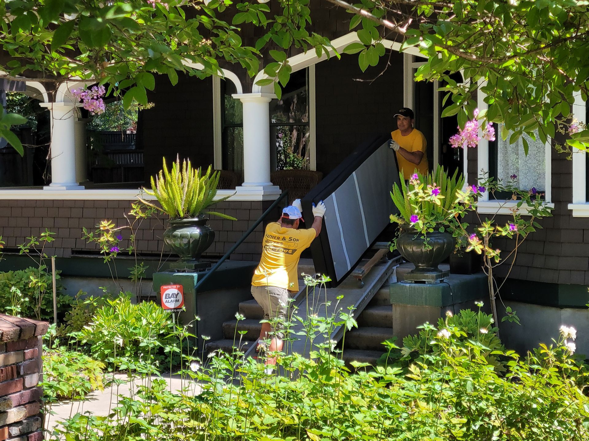 Person carrying a large flat-screen TV up front steps of a house with a porch and garden.