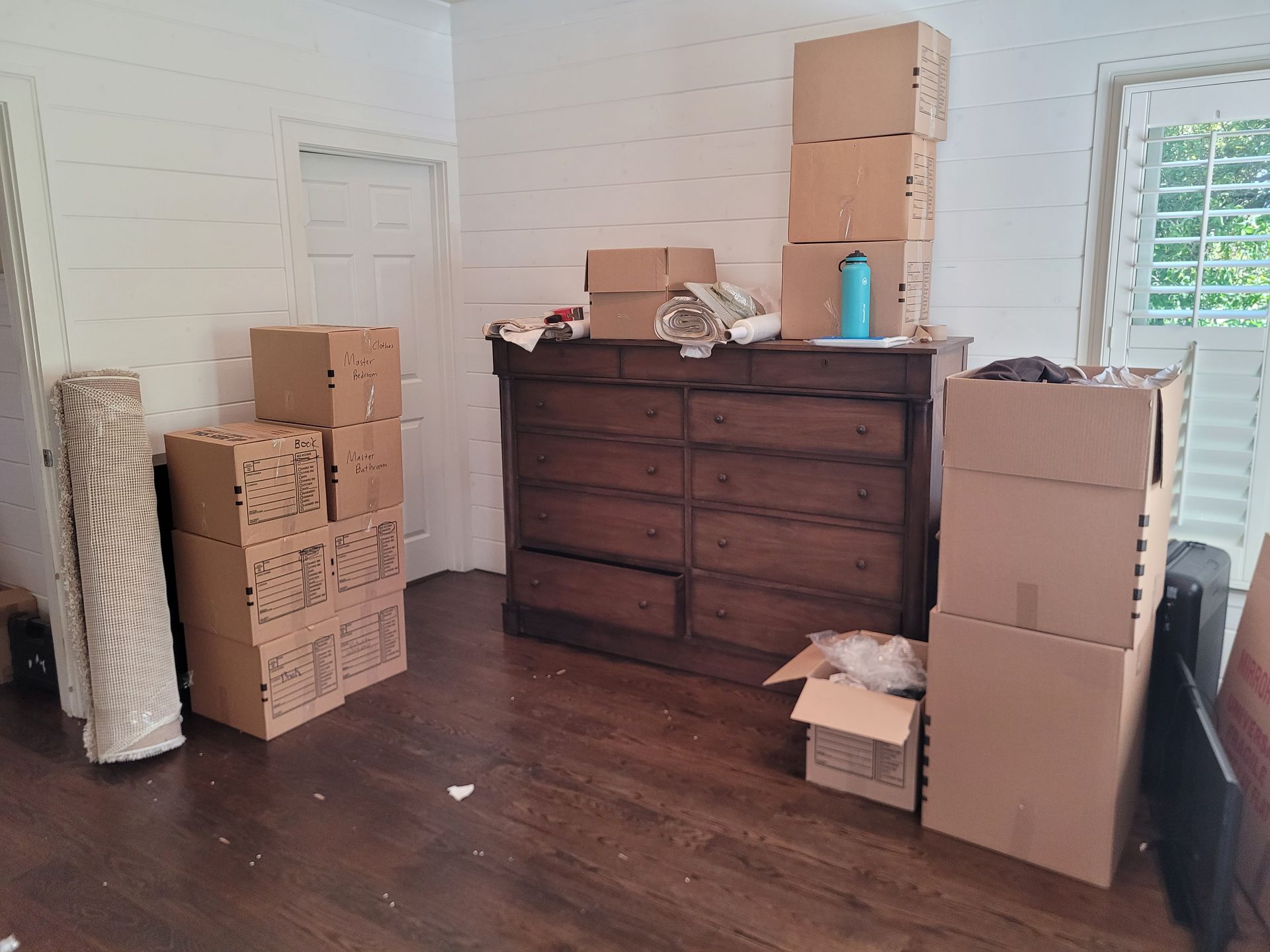 Unpacked moving boxes and a dresser in a mostly empty room with white walls and dark wood floors
