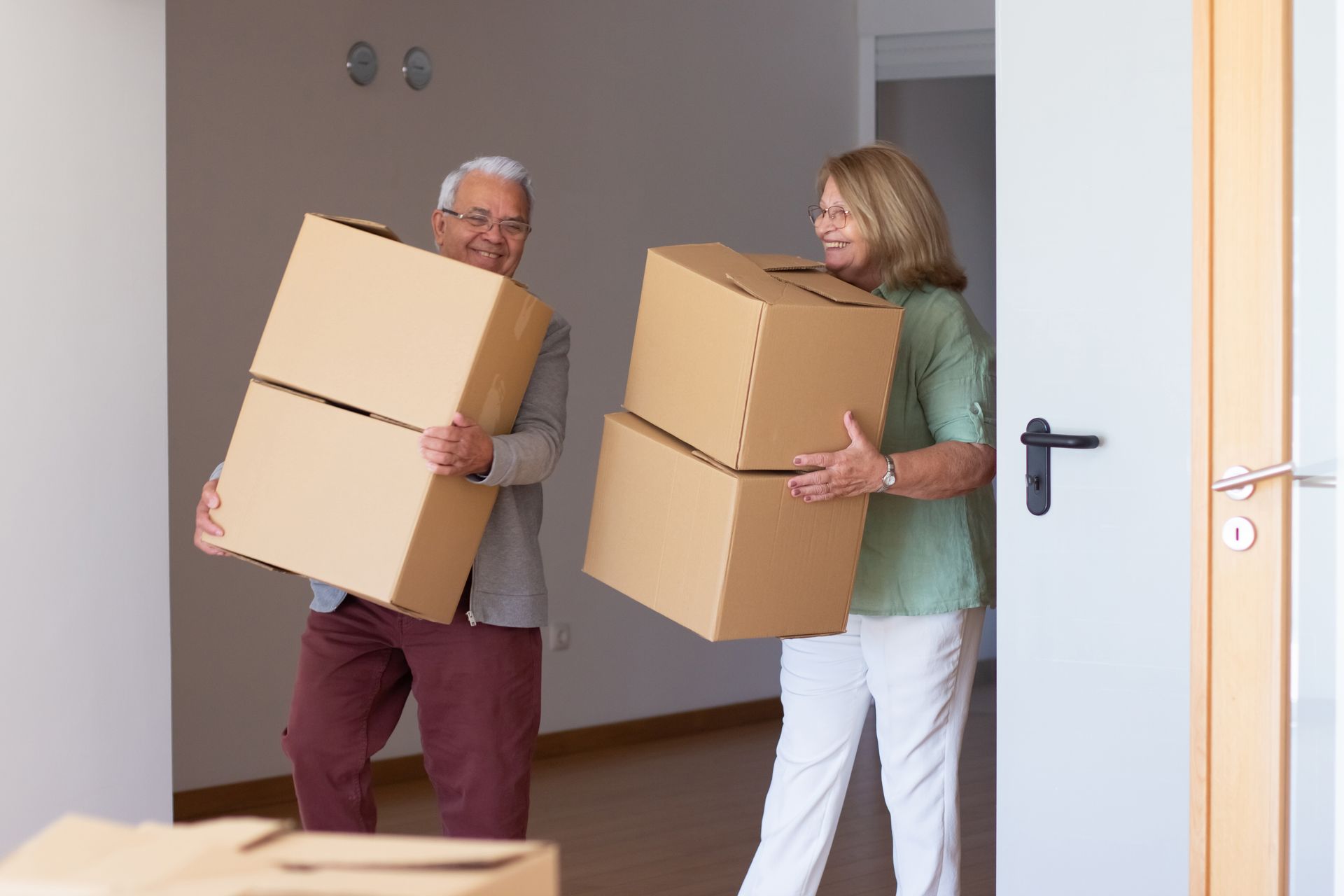 Two people carrying moving boxes into a new home, smiling.