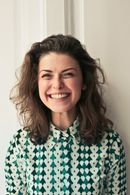 Woman with curly brown hair smiles broadly, wearing a green and white patterned shirt, in front of a white door.