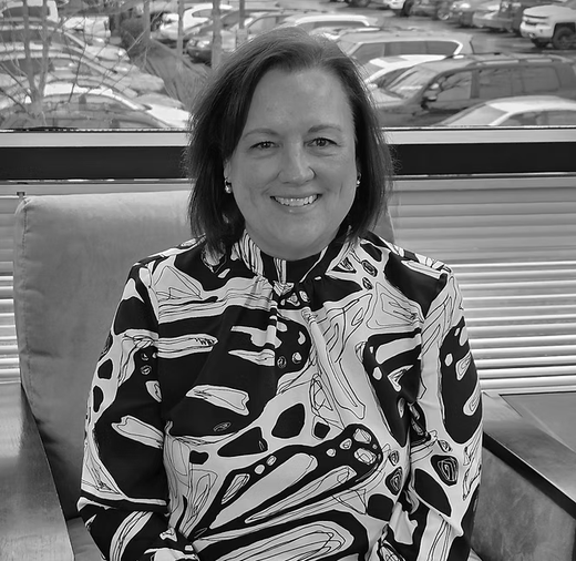 Smiling person seated indoors in a black-and-white patterned shirt, with cars visible through the window behind.