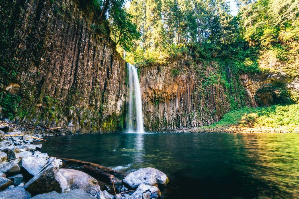 Tall waterfall dropping into a dark pool, surrounded by rocky cliffs and green forest under sunlight.
