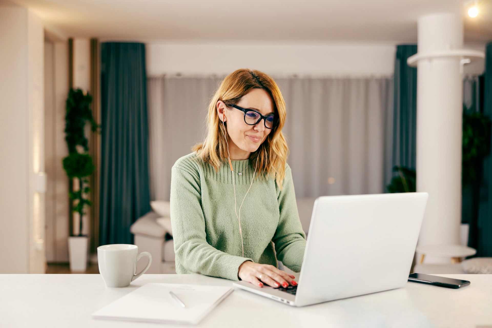 Person typing on a laptop at a home desk with a mug, notebook, and curtains in the background