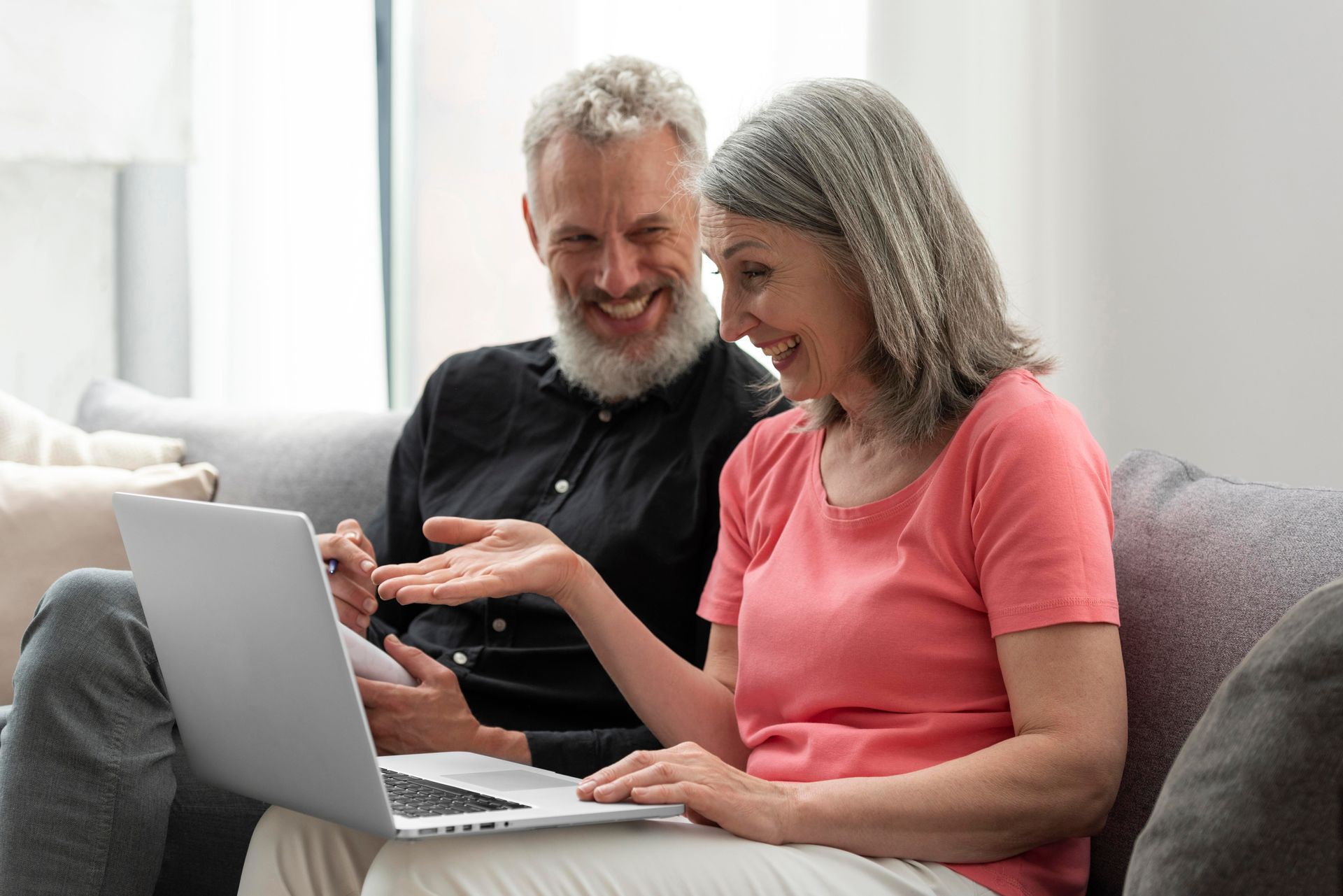 Two people smiling while using a laptop on a couch in a bright living room