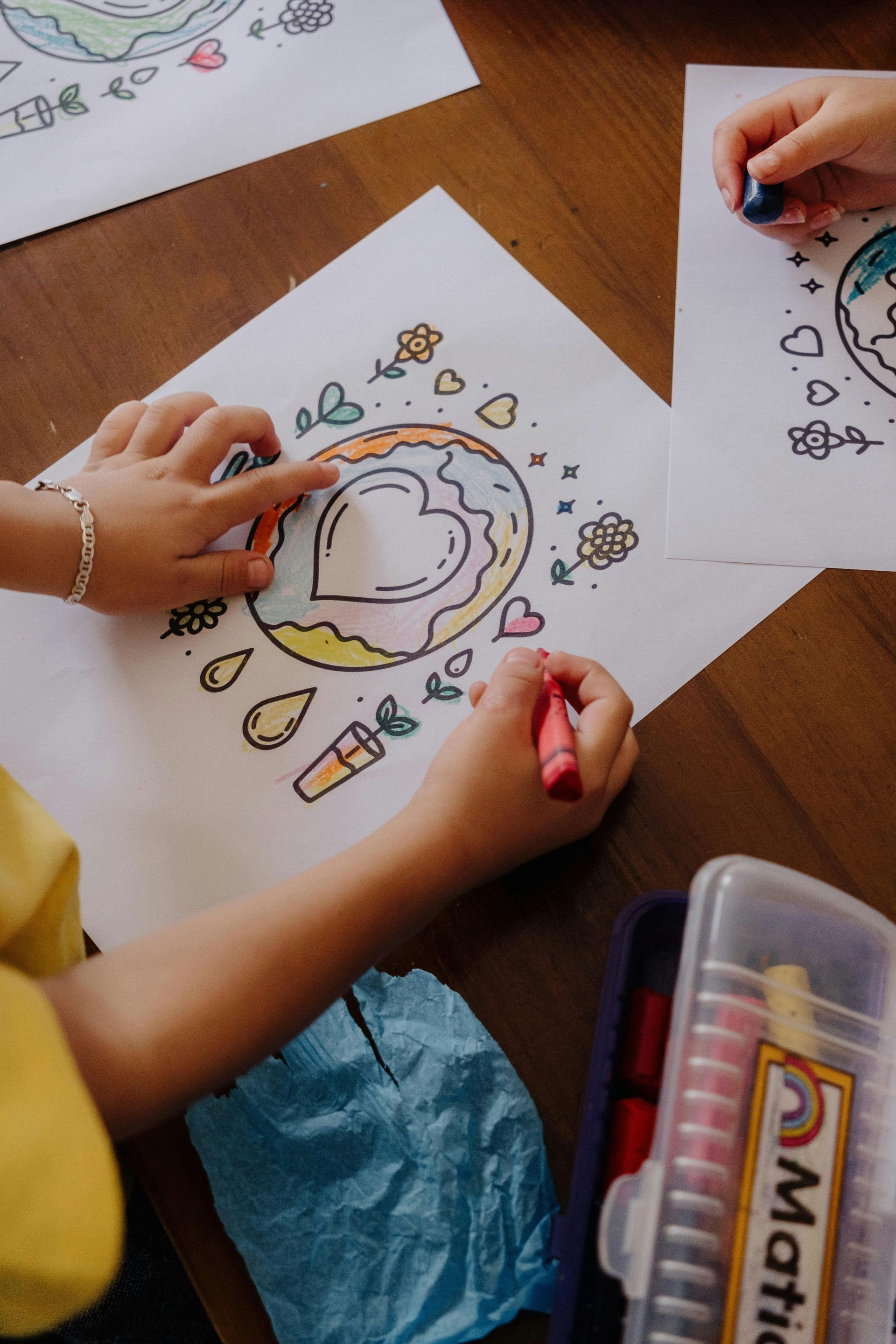 Children coloring a circular doodle on paper with crayons at a wooden table