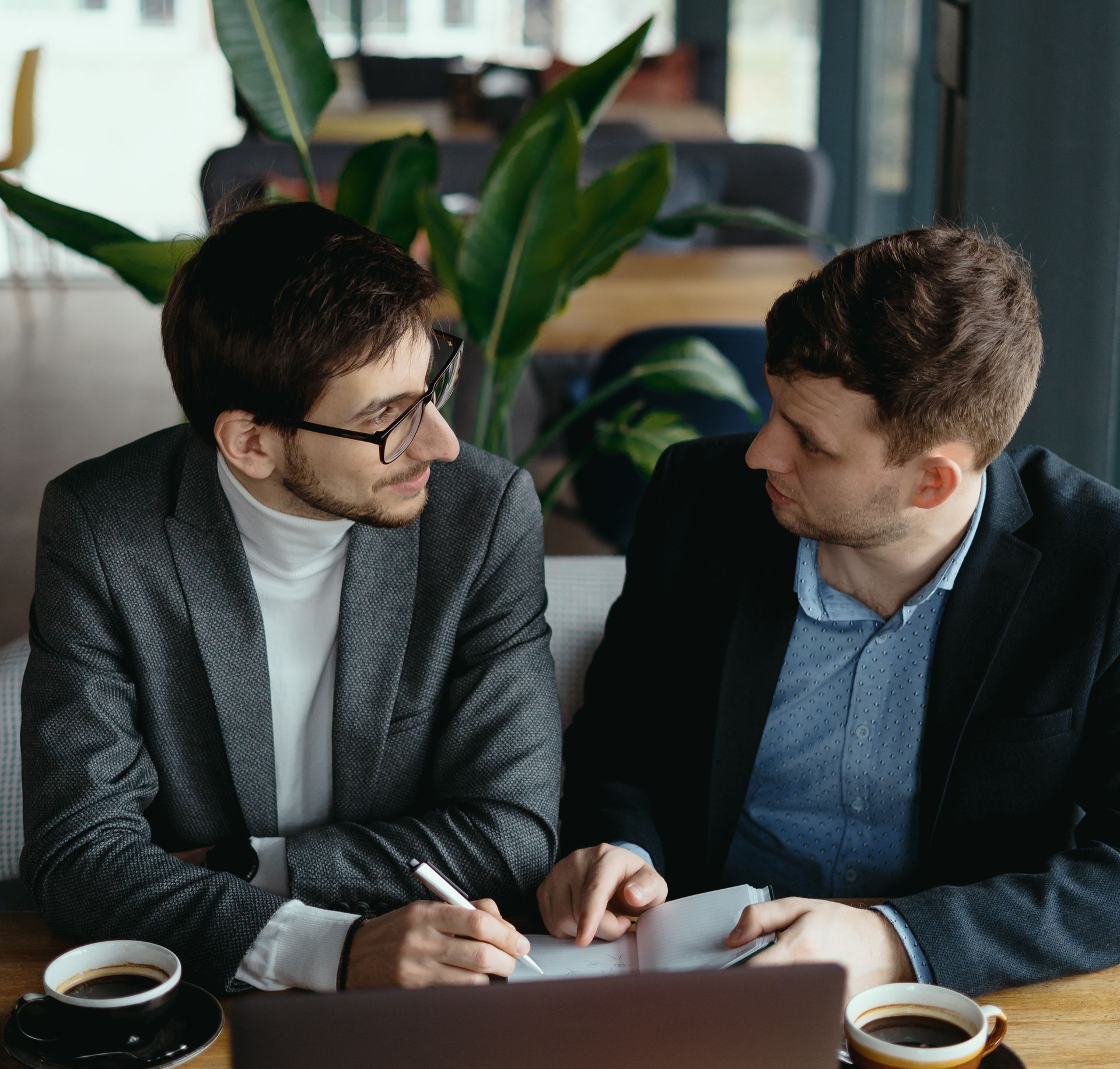 Two coworkers discussing notes at a laptop in a plant-filled office