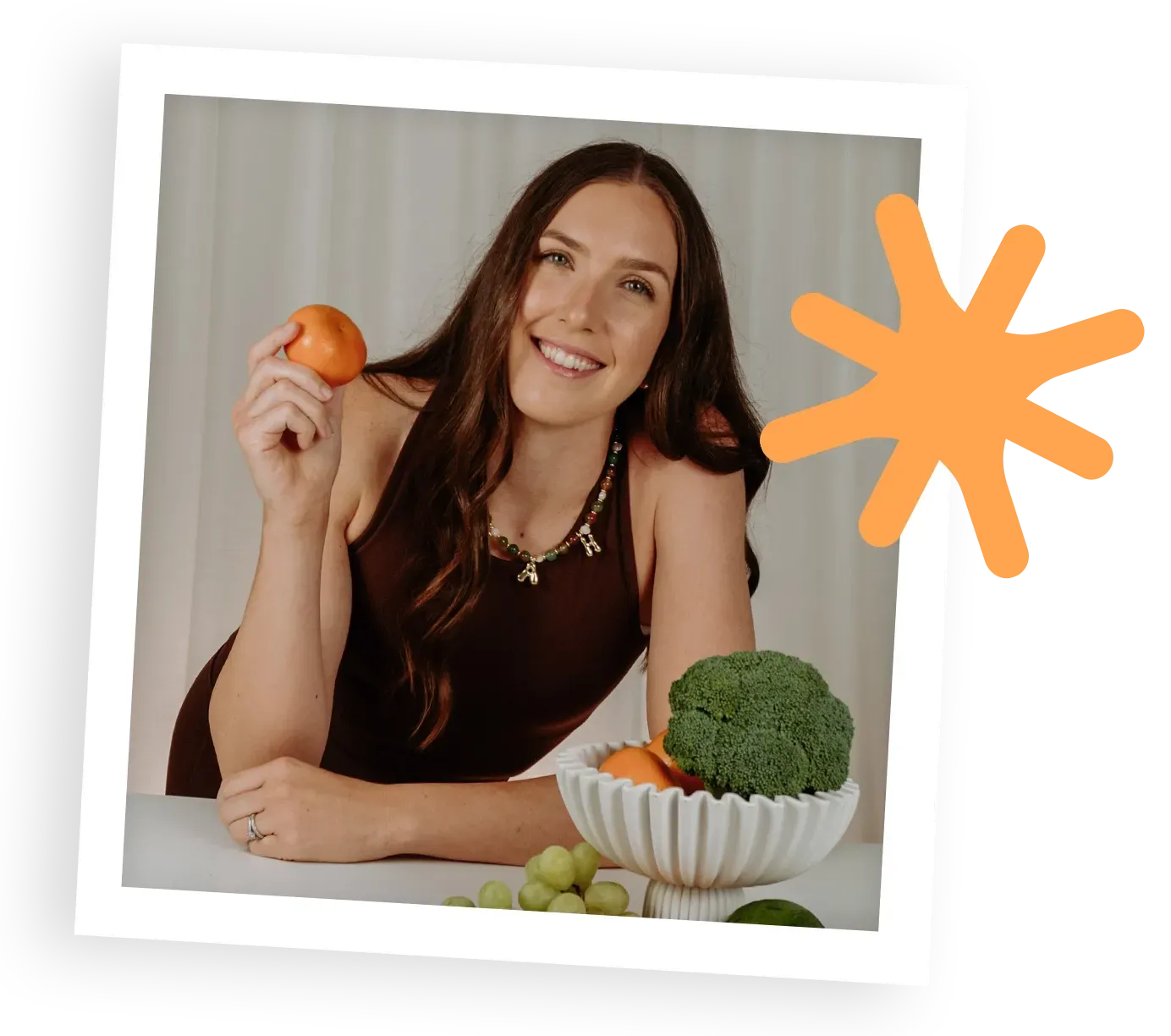 Zannetta holding an orange beside a bowl of fruit and broccoli