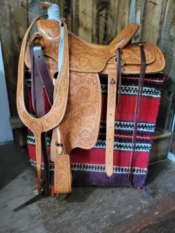 Brown leather saddle with straps hanging on a red-and-black woven blanket in a rustic shed