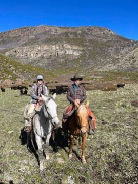 Two horseback riders on a grassy trail with mountains and blue sky in the background