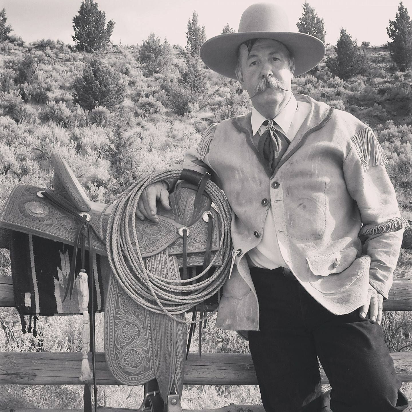 Black-and-white portrait of a cowboy in a hat leaning on a fence with coiled rope outdoors