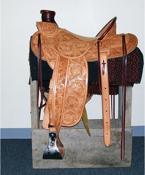 Ornate tan leather saddle on a stand, with straps and metal stirrup, displayed indoors