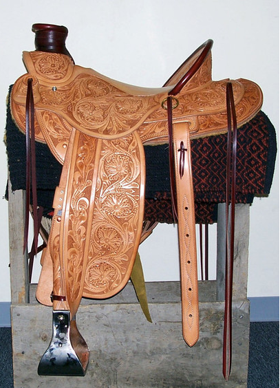 Tan leather Western saddle on a stand, with ornate tooling and dark blanket underneath