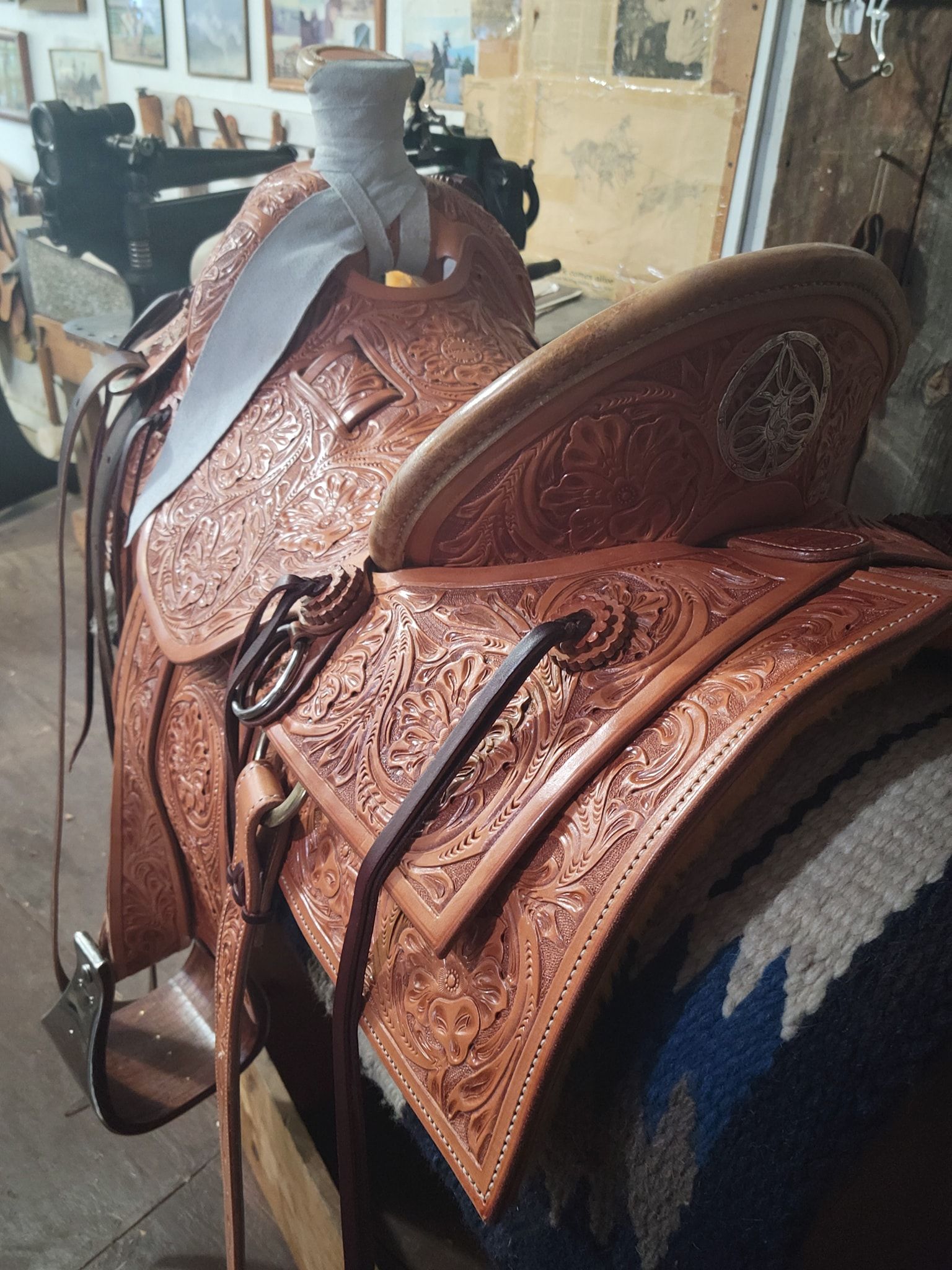 Brown leather saddle on a horse in a tack room, with a saddle blanket and wall art in the background