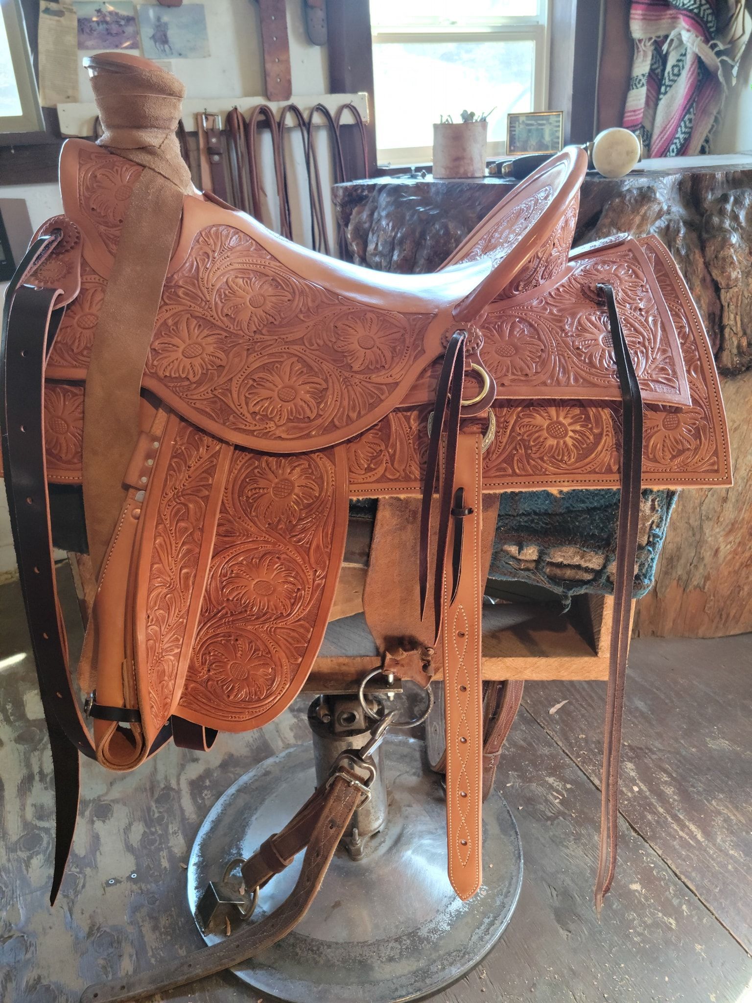 Brown leather saddle on a wooden stand in a workshop, with a bicycle wheel visible underneath.