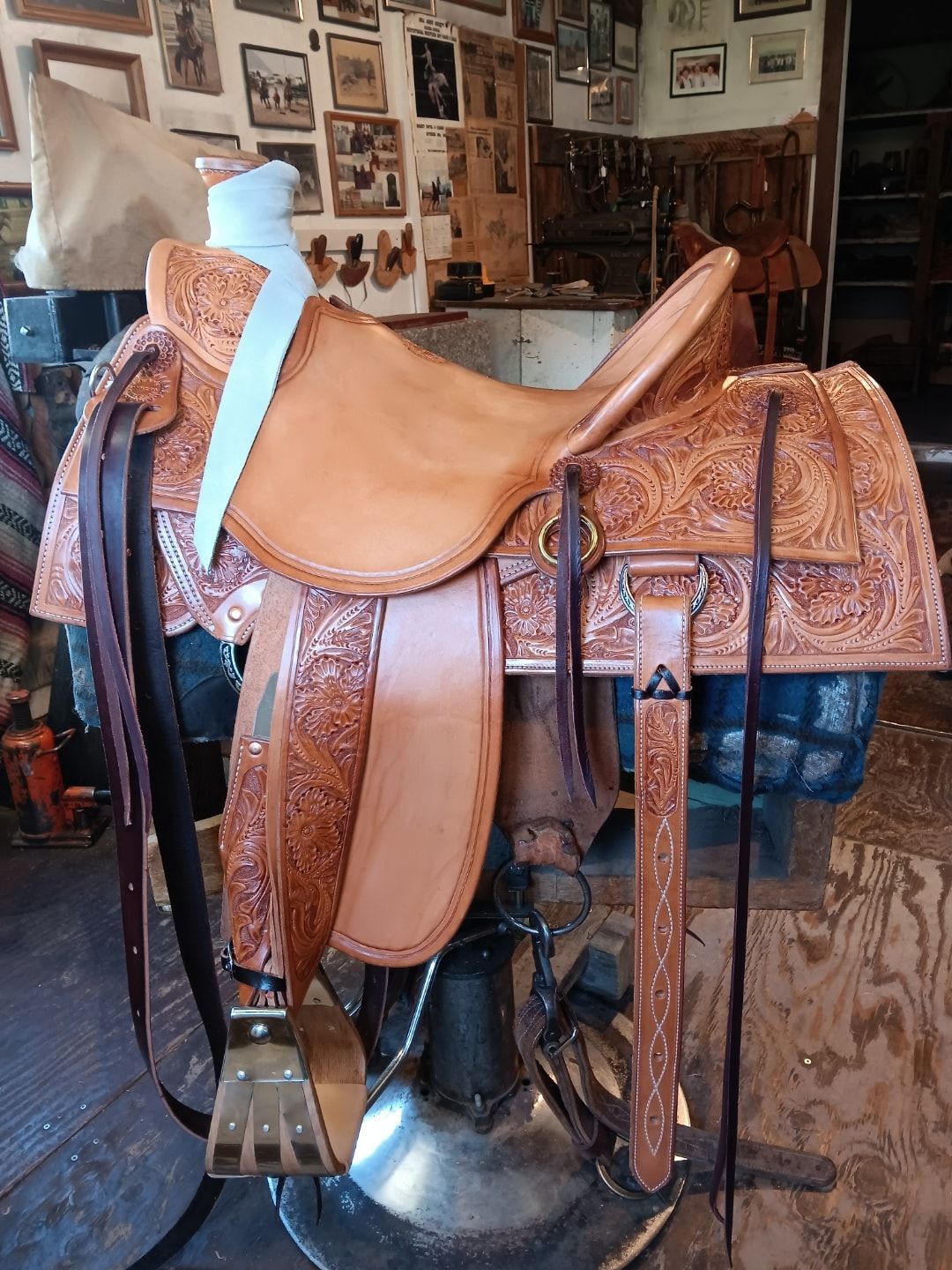 Brown leather horse saddle on a stand in a rustic tack room, with framed photos on the wall.