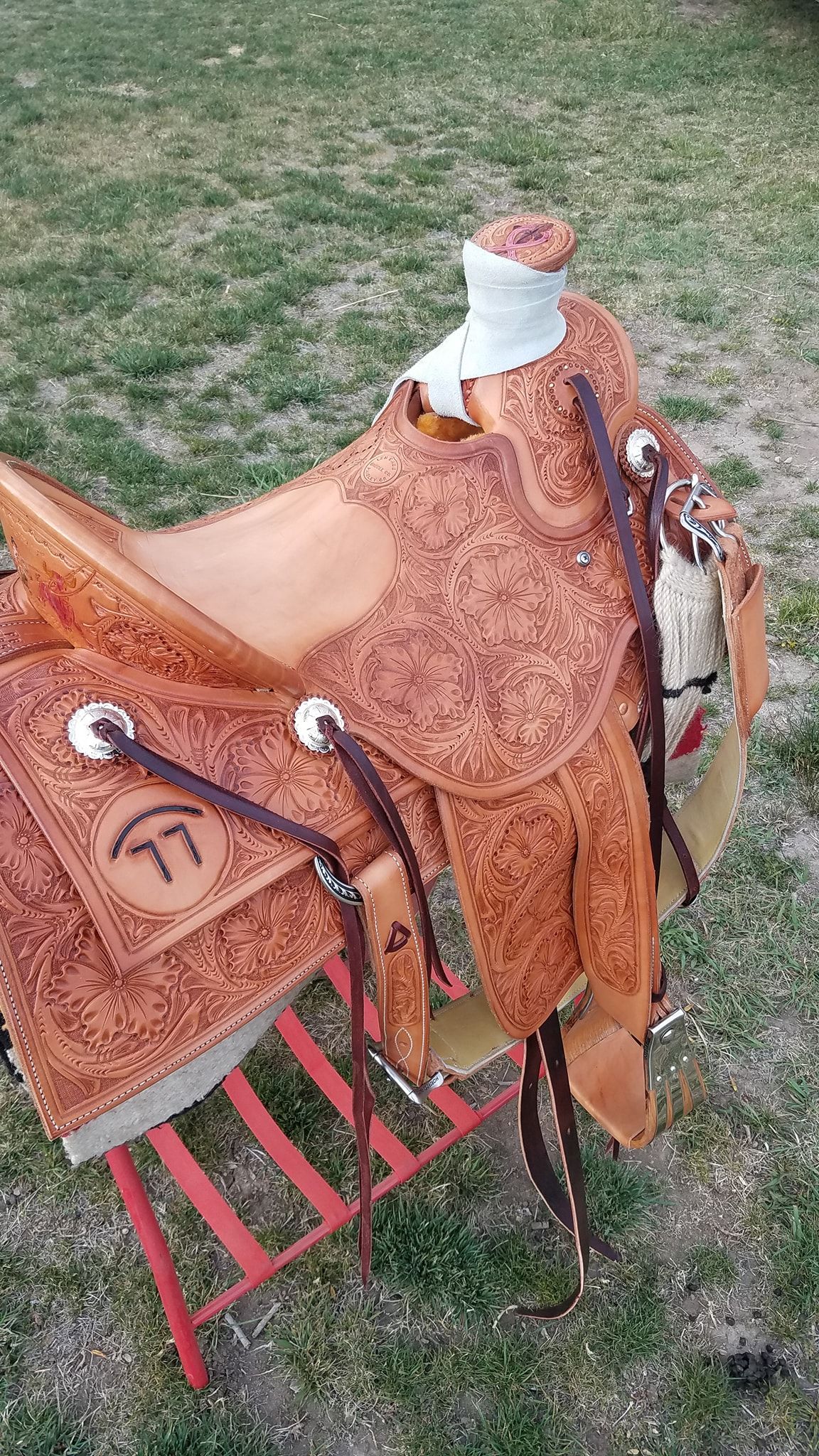 Brown leather saddle on a red rack outdoors on grass, with saddle pads and straps attached