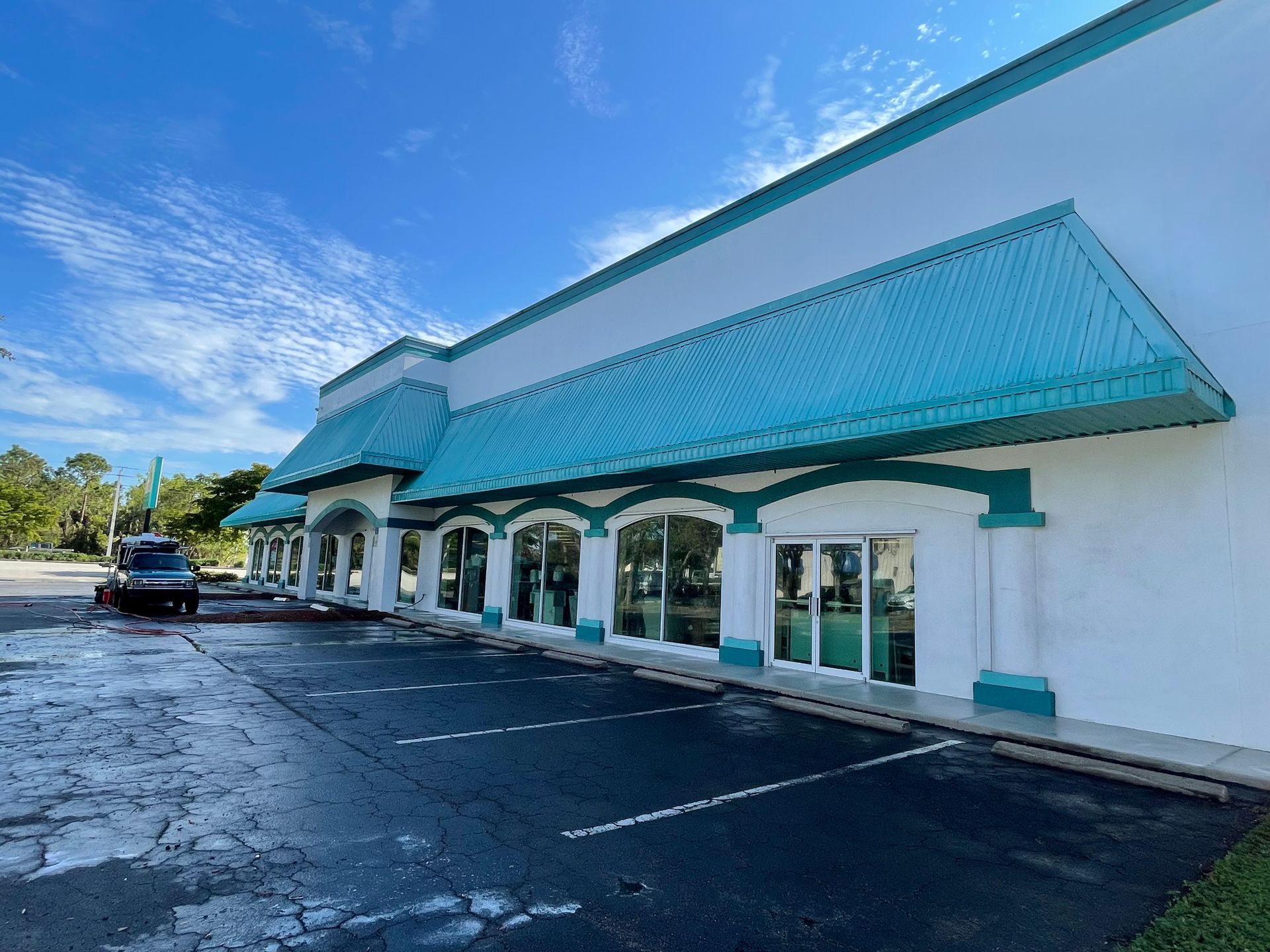 A commercial building with a white facade and teal metal awnings above storefront windows, beside an empty parking lot.