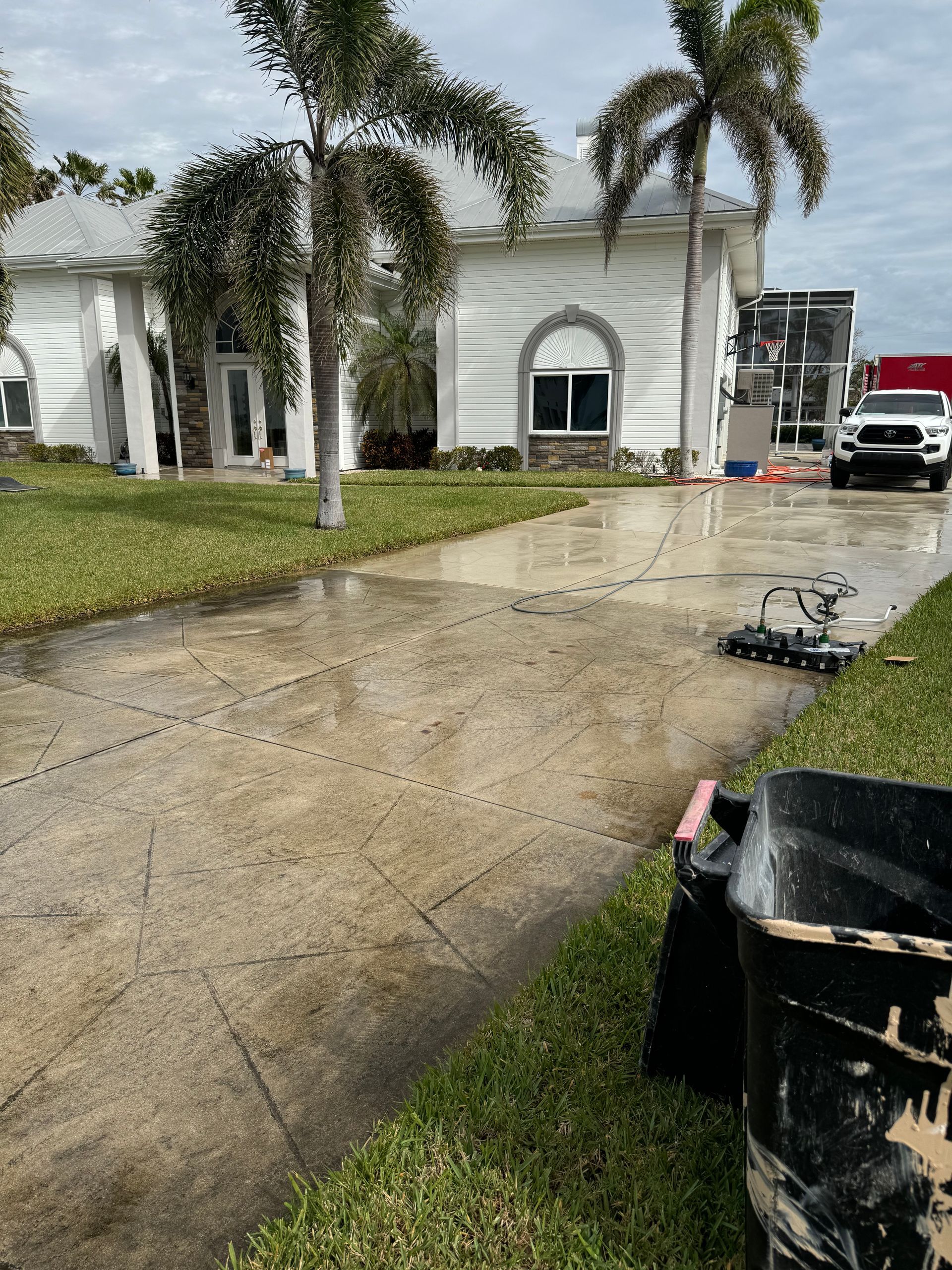 A pressure-washed driveway with a surface cleaner sitting in the middle, next to a white house with palm trees.