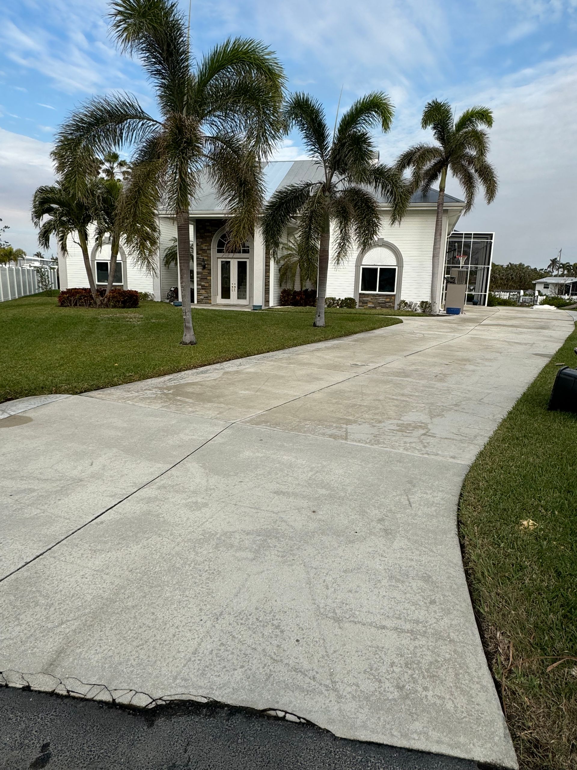 A white, single-story house with a long concrete driveway surrounded by palm trees and green lawn under a blue sky.