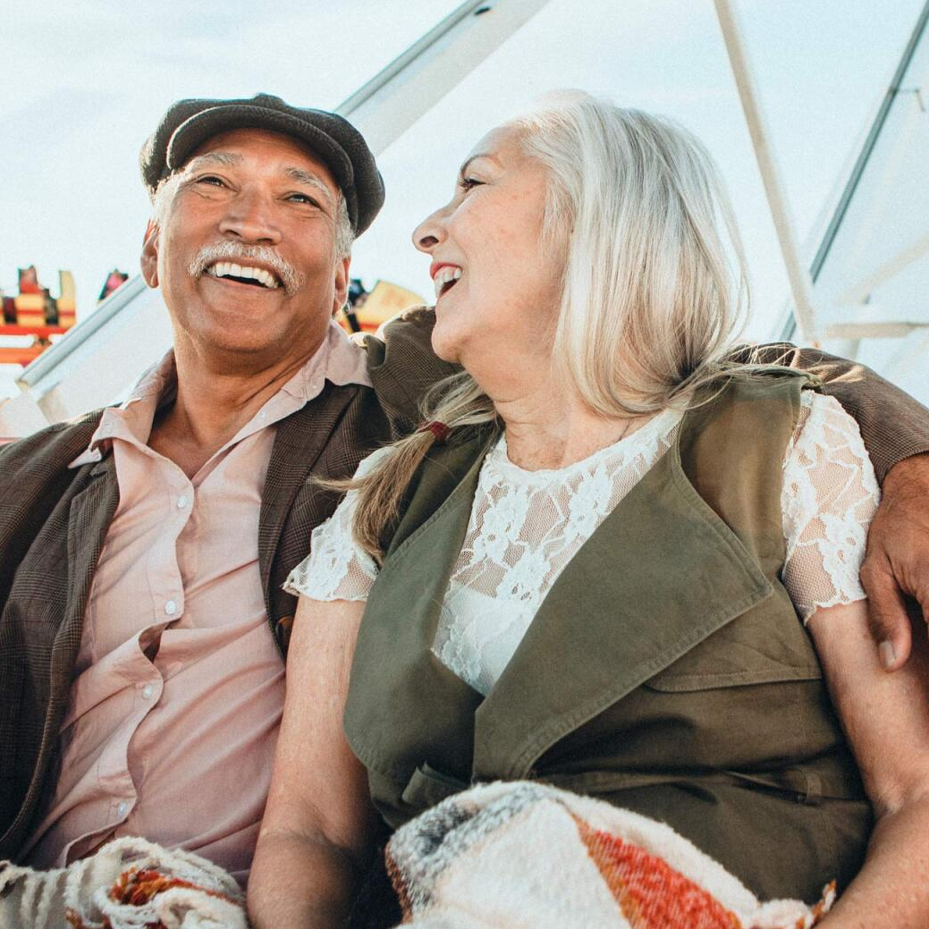 Two smiling people sitting together in a bright outdoor setting, laughing and looking at each other