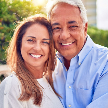Smiling couple standing outdoors in bright sunlight, with greenery softly blurred in the background