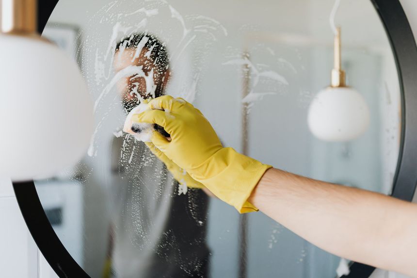Gloved hand cleaning a mirror with spray cleaner, wiping away smudges on the glass.