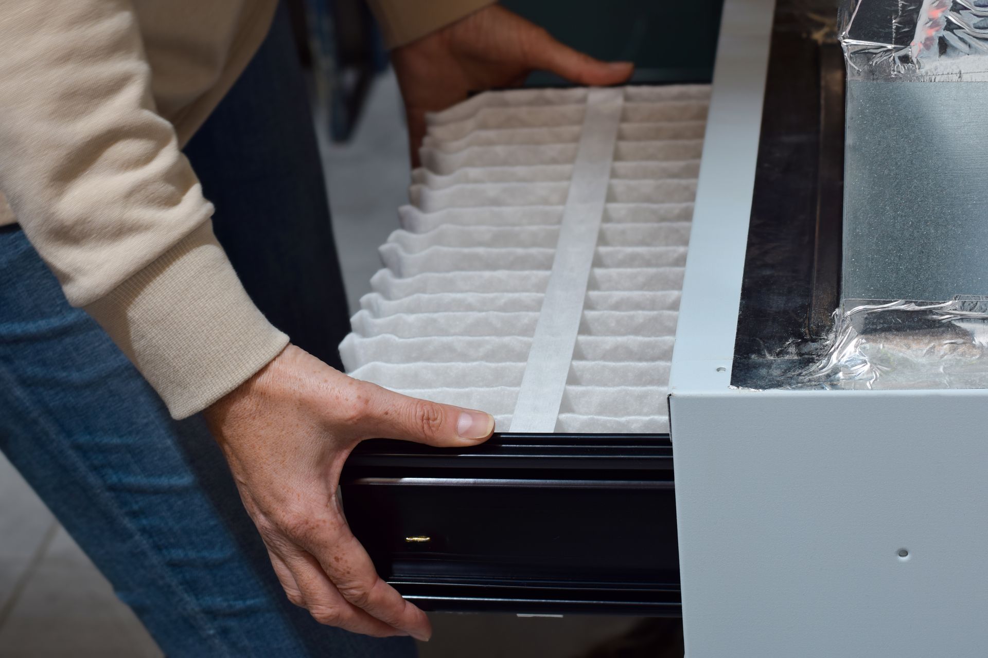Person replacing a furnace filter. Hands holding a new, white pleated filter near a metal unit.