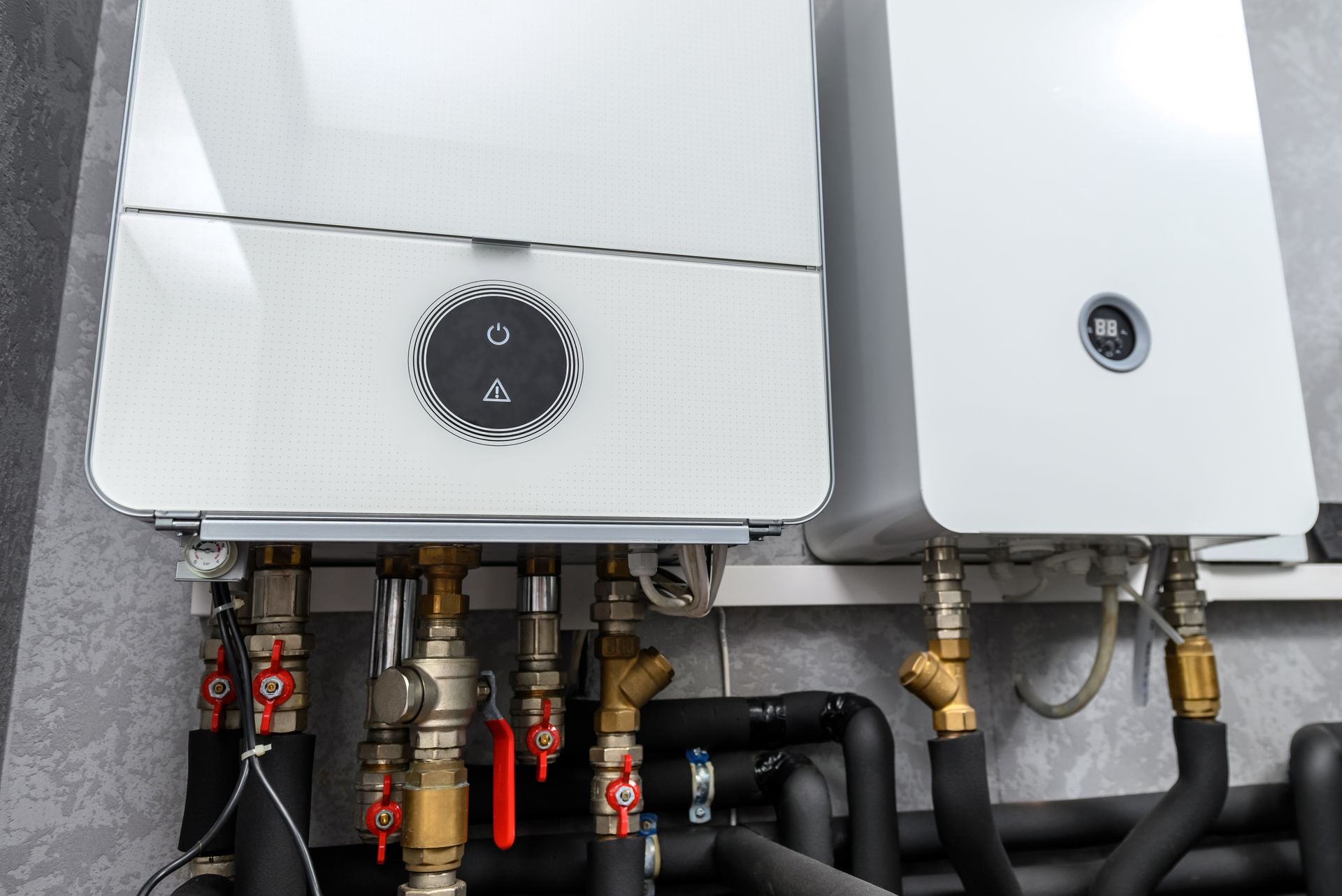 Two white heating boilers with attached pipes and valves against a grey wall.