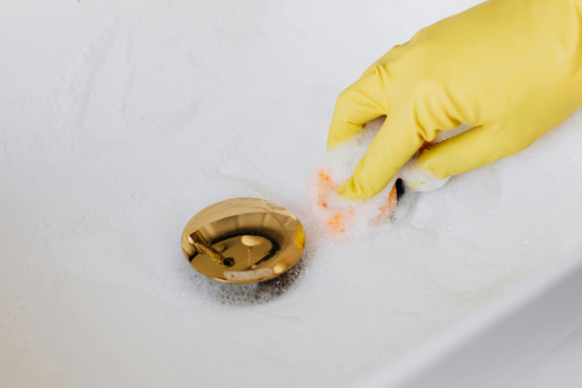 A gloved hand cleans a white sink with soap suds near a gold drain.