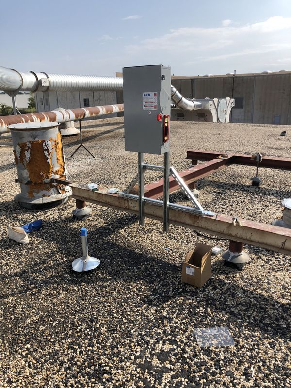 Gray electrical box on a rooftop, supported by metal legs and a beam. A box and pipe are nearby.