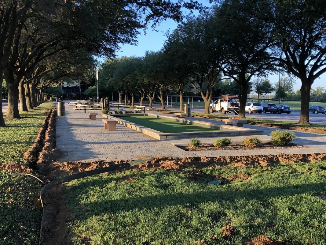 A park with bocce ball courts and benches, lined with trees, near a parking lot.