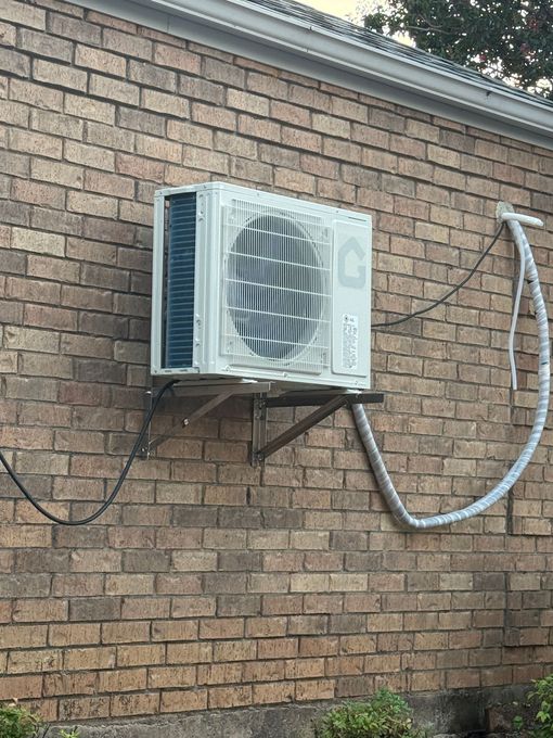 Exterior air conditioning unit mounted on a brick wall with visible power and refrigerant lines