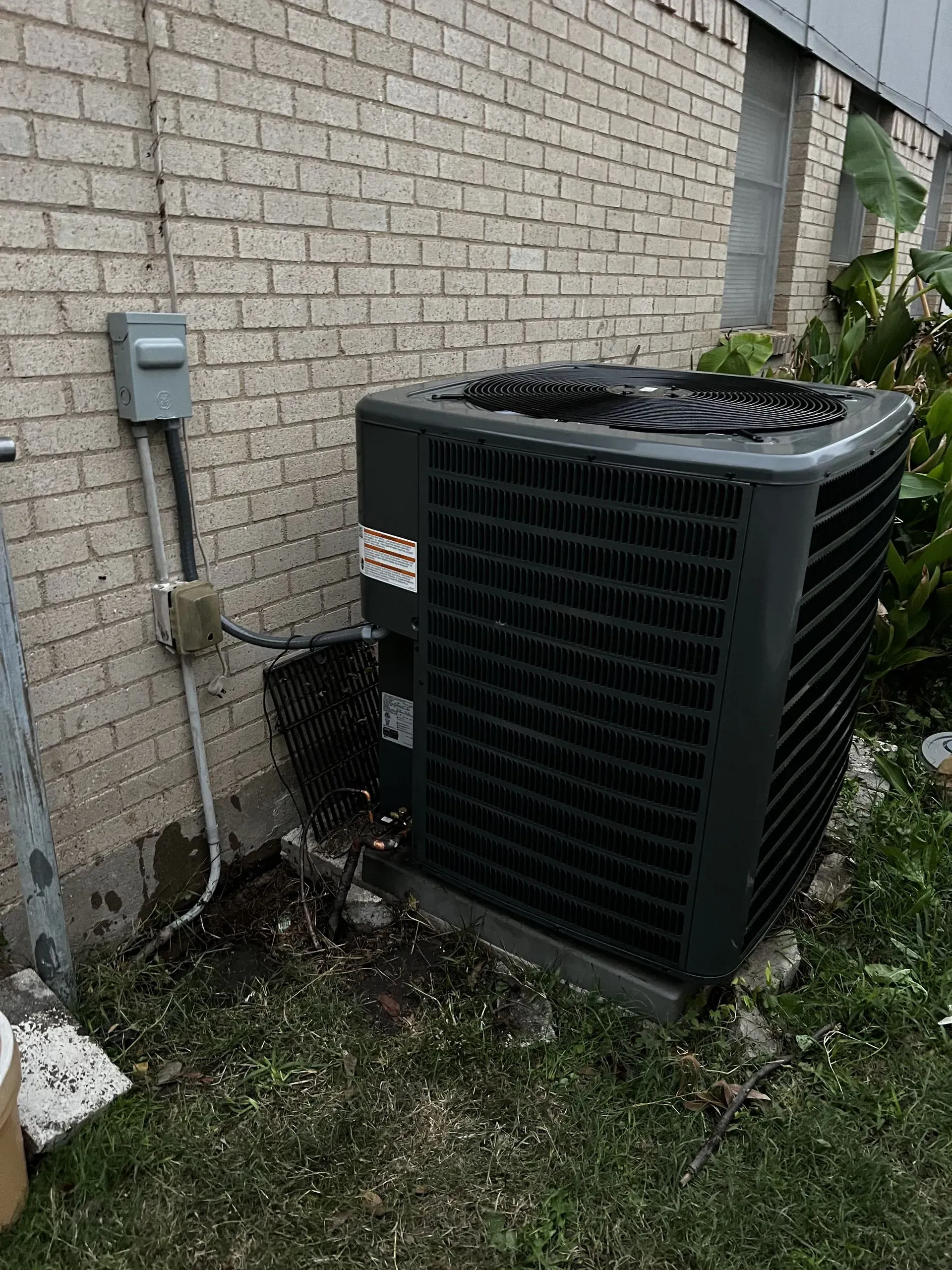 Outdoor air conditioner unit beside a brick house wall with electrical meter and conduit.