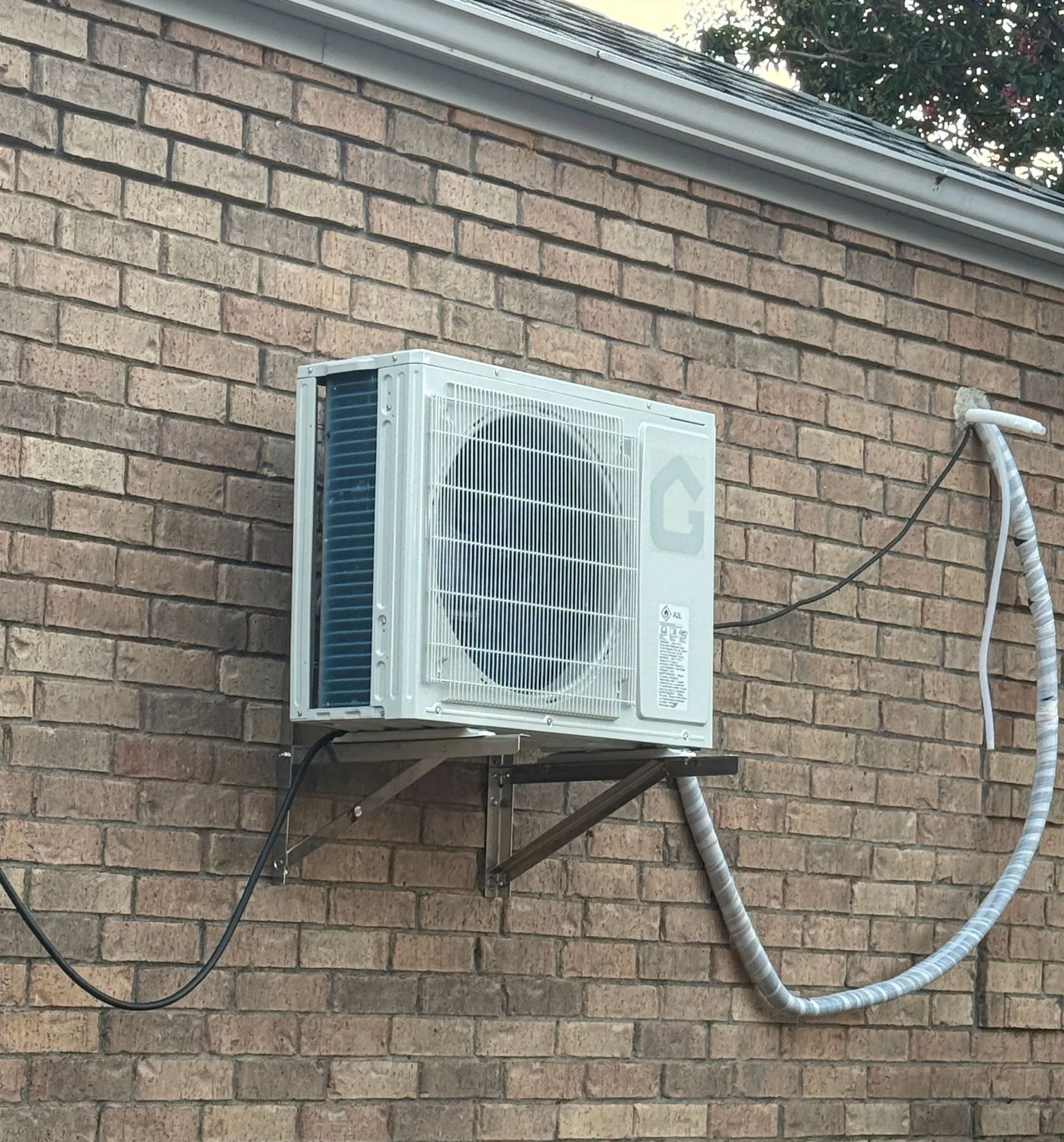 White wall-mounted air conditioner on a tan brick wall with exposed hoses and cables.