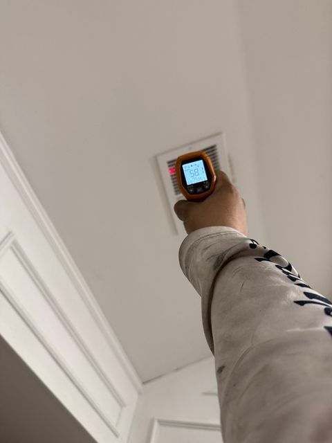 Hand holding a handheld stud finder up to a ceiling near a vent in a white room