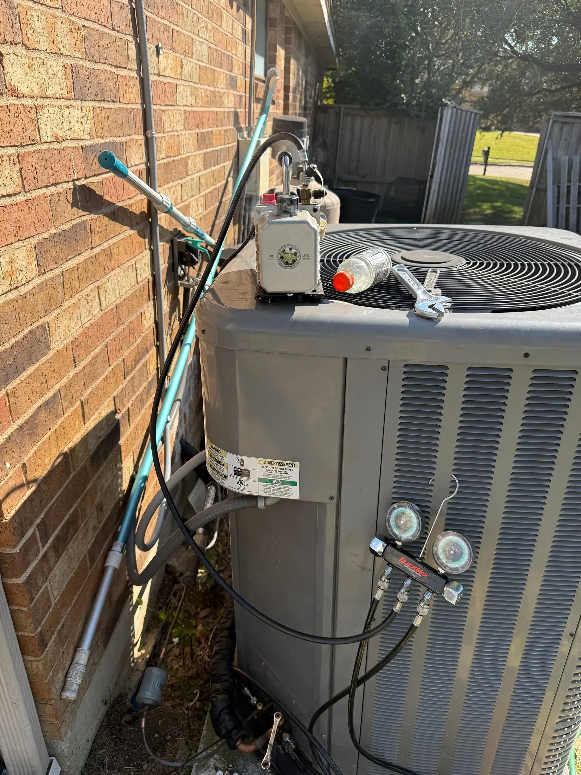Outdoor HVAC unit beside a brick wall with refrigerant gauges and hoses attached