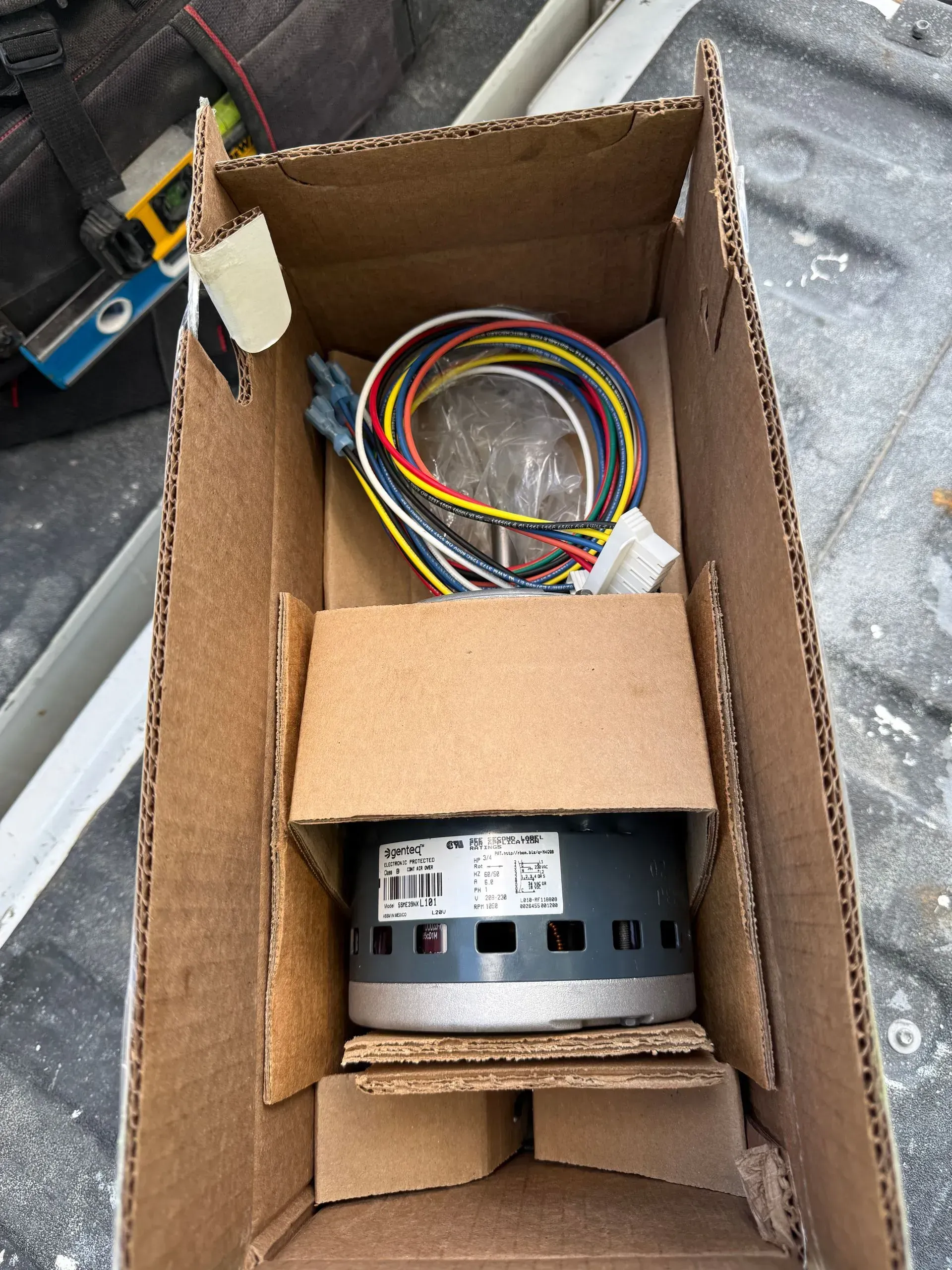 Open cardboard box holding coiled multicolored wires and a metal component in a car trunk