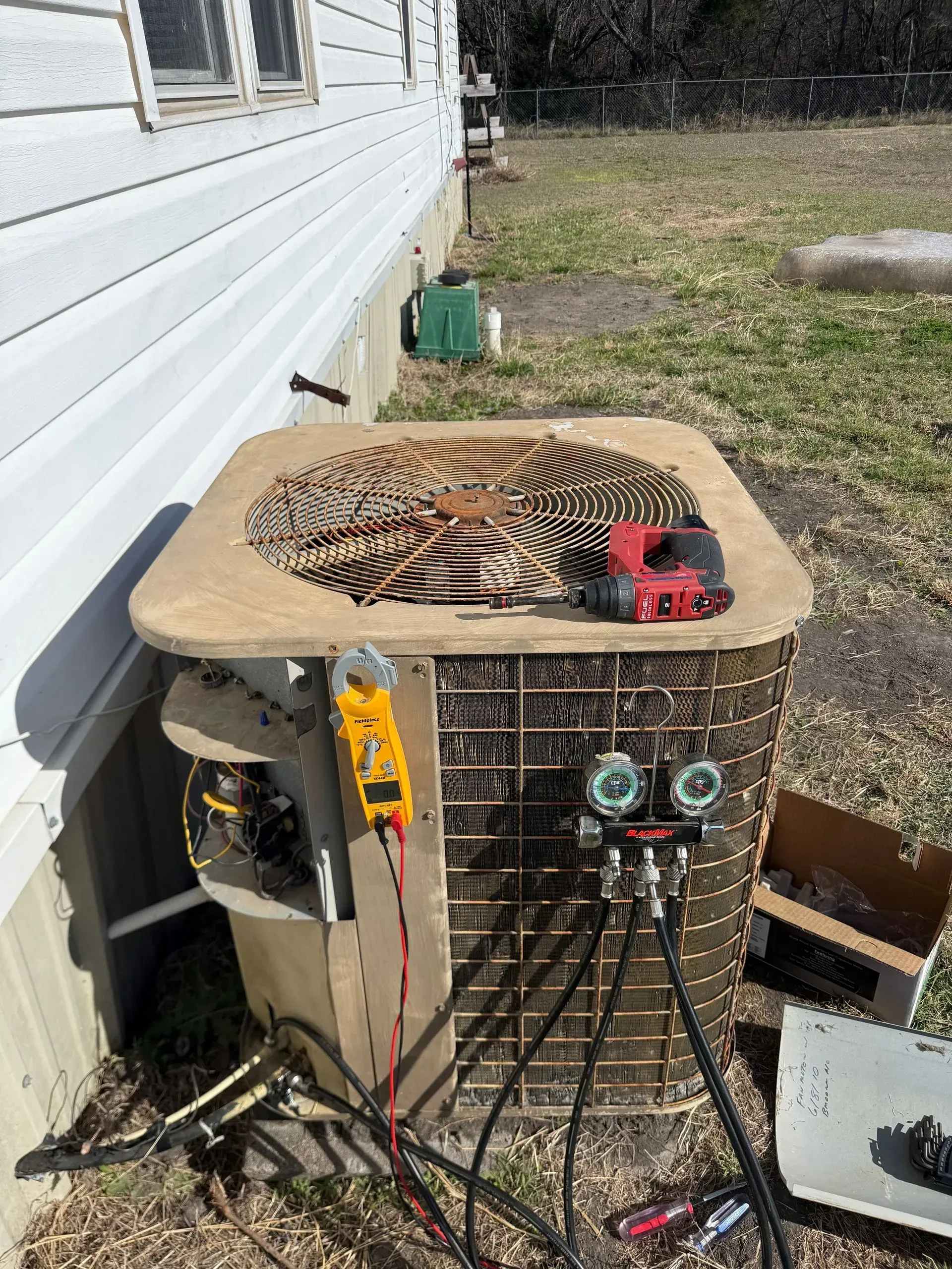 Outdoor HVAC unit beside a white house, with tools and cables hanging from it and debris on top