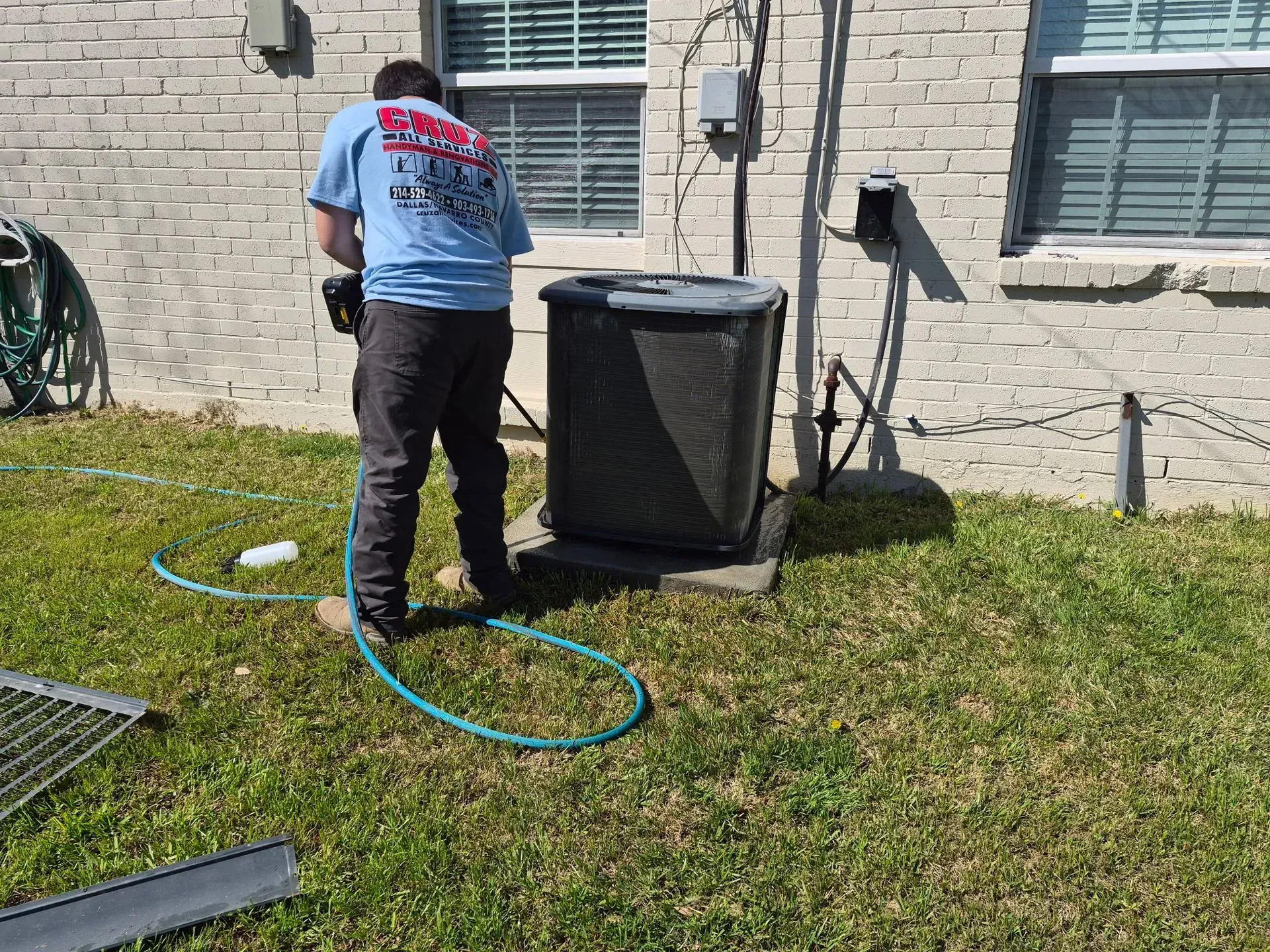 Person pressure-washing an outdoor HVAC unit beside a brick house with a hose on the lawn